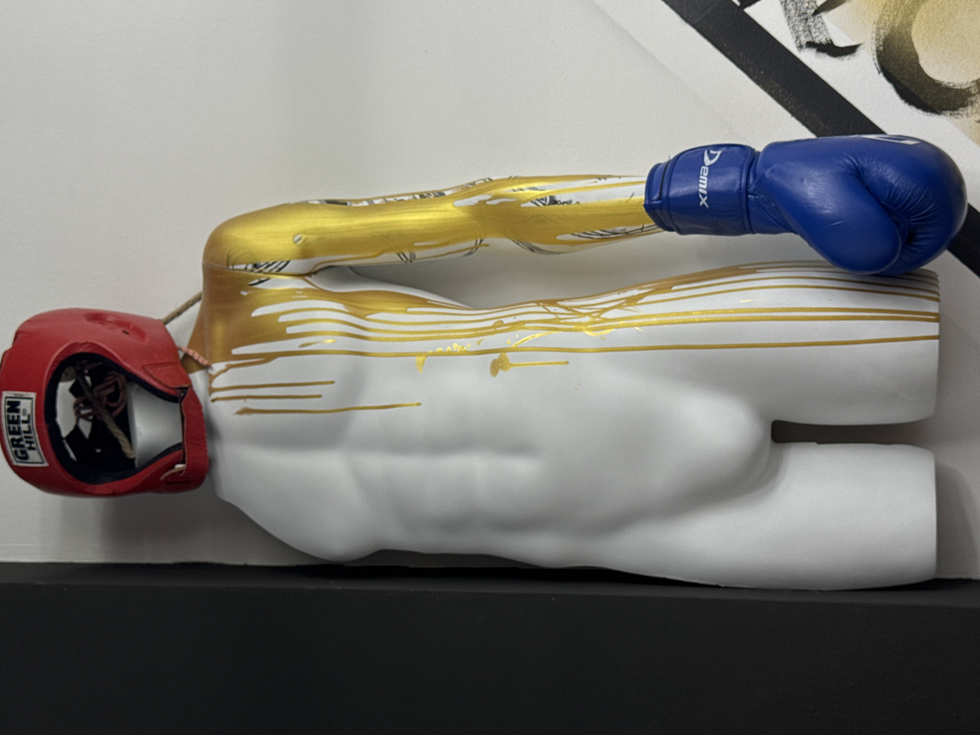 The scene depicts a white, male-form mannequin torso, headless and legless, displayed vertically. Gold paint drips are evident, running down the front of the torso and extensively covering the right arm, which also features dark linear markings resembling tattoos beneath the gold. A red boxing headgear, branded "GREEN HILL" in white lettering, rests atop the mannequin's neck opening. A single blue boxing glove, bearing the white "DEMIX" logo, is attached to the mannequin's right arm, positioned as a closed fist. The mannequin is situated in an interior corner, with a dark, possibly black, wall segment on its left and a white wall on its right. A partial view of an abstract artwork with black and gold elements is visible on the lower right background. The image provides no visual information to confirm the location in Chaikovsky, Russia.