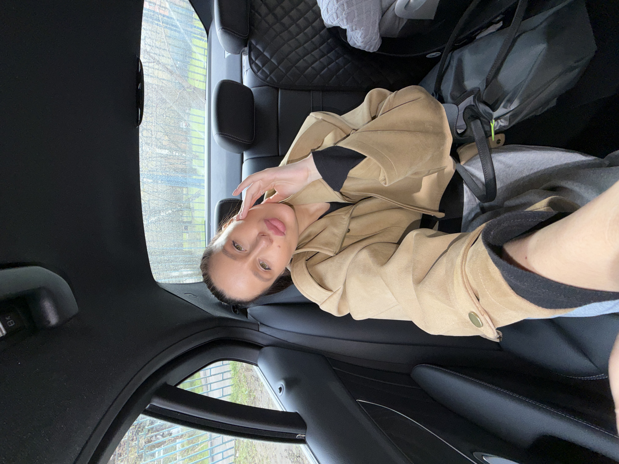 A person is lying in the back seat of a car. The person is wearing a tan jacket and black shirt. A gray bag is visible near the headrest of the seat. Outside the car window, there's a blurred view of a green area, possibly in Khimki, Russia. The car's interior is primarily black leather.
