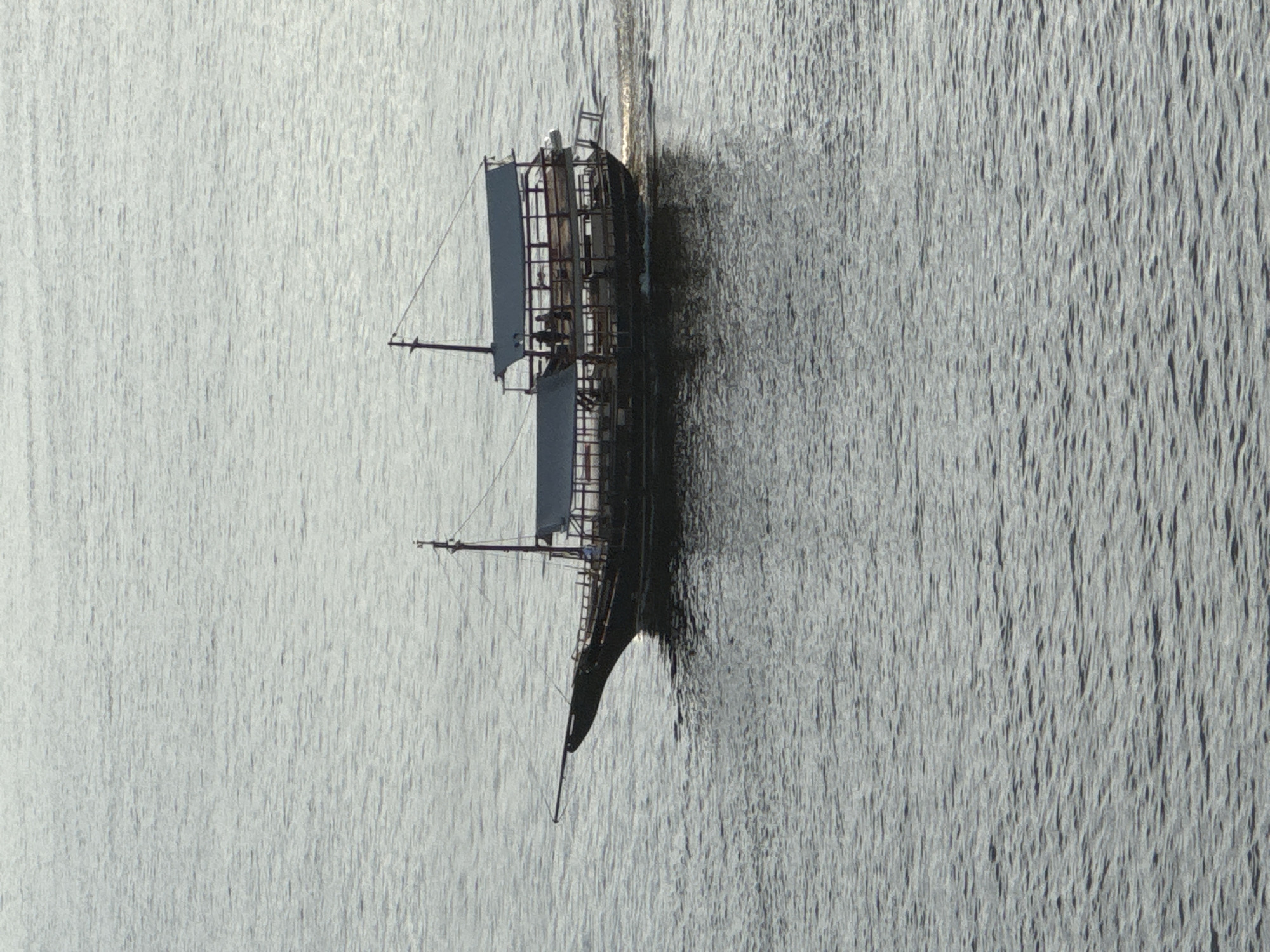 A dark, wooden boat with multiple decks and two masts sails on the water. The boat is moving from left to right, creating a wake behind it. There are several figures visible on the decks of the boat. The water is rippled and appears to be a muted grey color, reflecting the overcast sky. The boat is positioned in the middle ground of the image. The scene is likely set on Guanabara Bay, which borders the city of Niterói, Brazil.