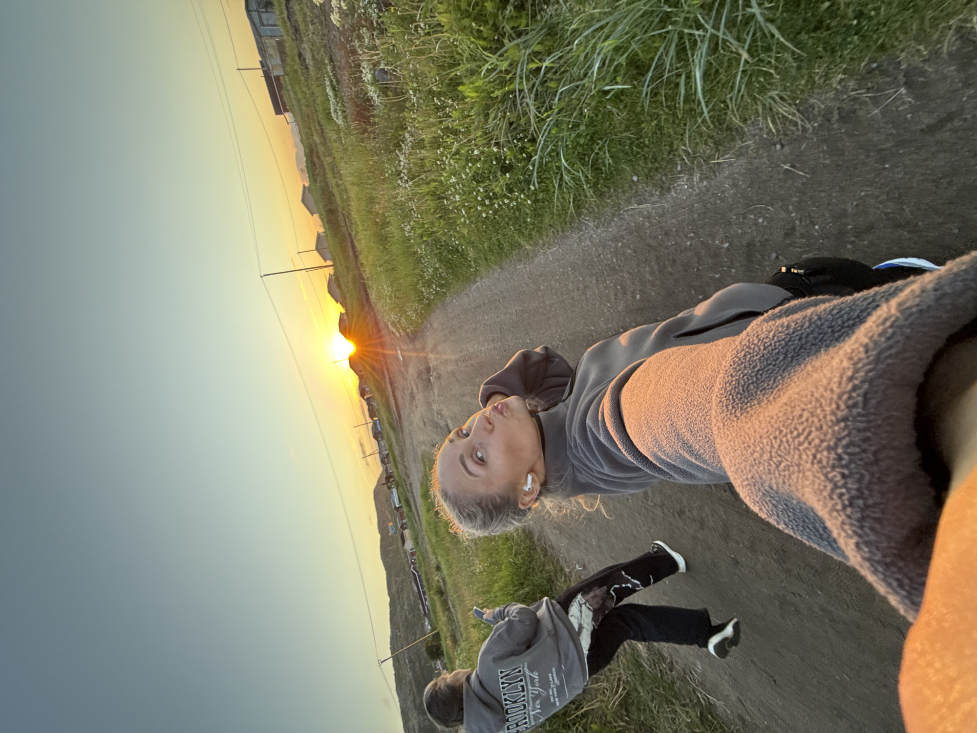 In Teriberka, Russia, at sunset, two individuals are captured in a selfie-style photograph on a dirt road lined with lush green grass and small white wildflowers. The sun is low on the horizon, casting a warm glow and creating a sunburst effect. In the foreground, the arm and shoulder of the person taking the photo are visible, wearing a gray fleece jacket. Adjacent to them, a woman with blonde hair tied up in a bun is posing for the camera with puckered lips and wearing white earbuds and a gray fleece. Further down the road, another individual is walking away from the camera, seen from the back, wearing a gray hoodie with "BROOKLYN NEW YORK" printed on it and black pants with a camouflage pattern. In the background, several small houses and power lines are visible, nestled against rolling hills under a clear sky transitioning from blue to yellow.