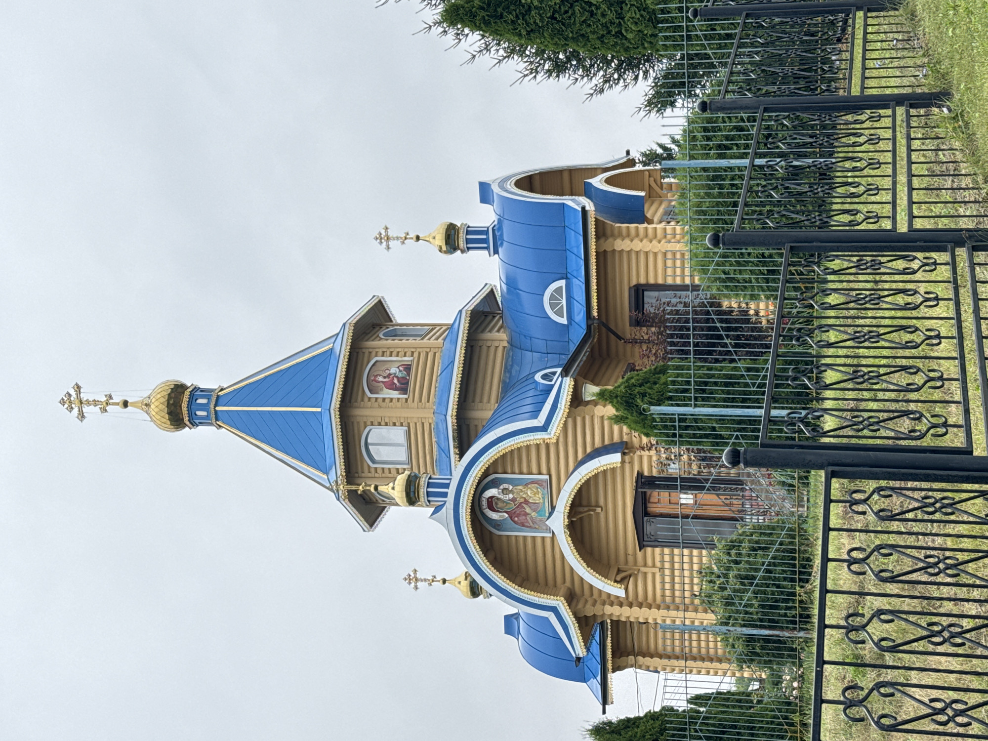 The image shows a light brown wooden church with blue roofs and gold crosses. The church has several domes and is adorned with religious iconography on its facade. A black metal fence runs along the foreground of the image, separating the viewer from the church grounds. The church appears to be located in a rural or suburban setting with trees and greenery visible around it. The sky is overcast. The specific location mentioned is Tashla, Russia.
