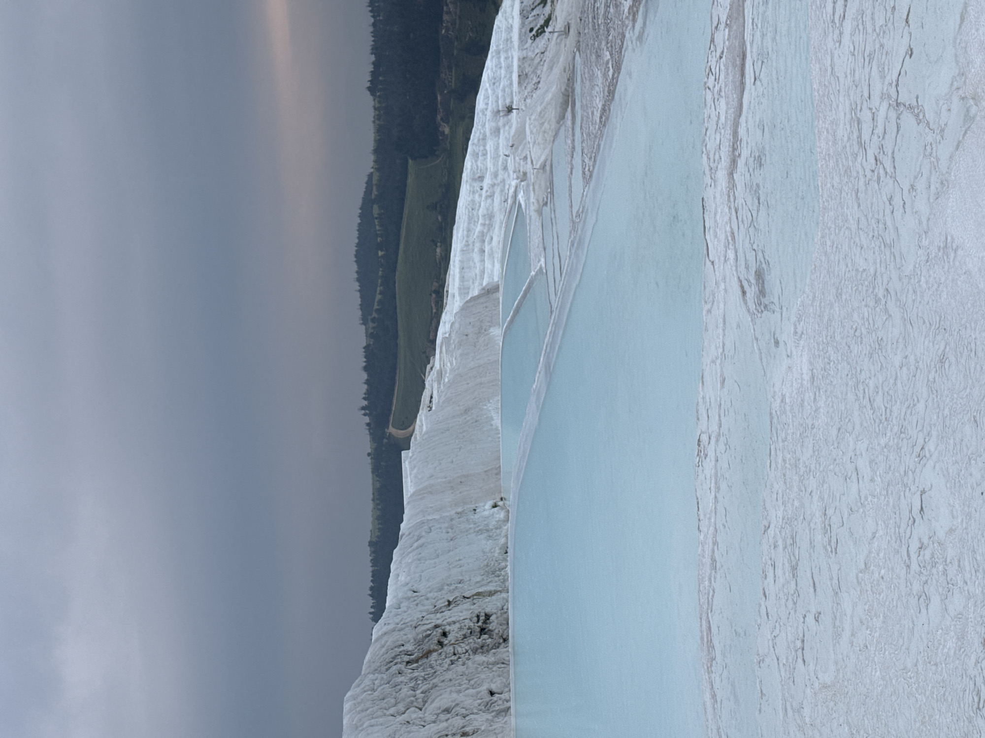 The image depicts the Pamukkale travertines in Turkey, showcasing the white, calcium carbonate terraces with light blue pools of mineral-rich water. In the background, a forest and overcast sky add to the scene. People are walking and exploring the area.