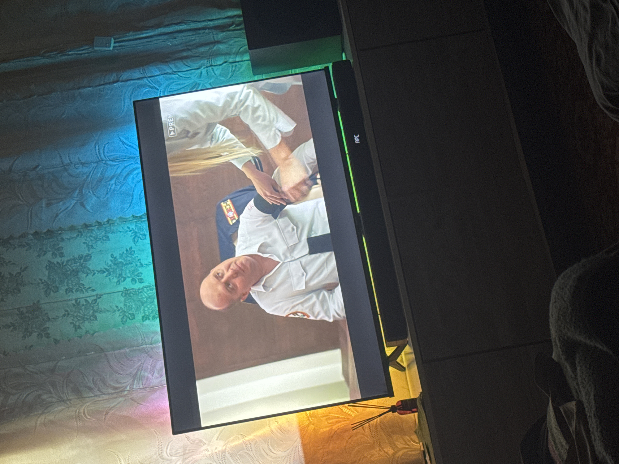 A television displays a scene with two people. One person wearing a white uniform is performing CPR on another person. The setting appears to be indoors. The location is not discernible from the image.