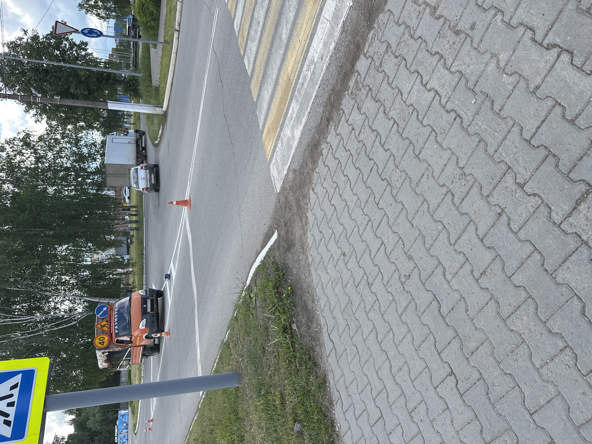 This media file captures an outdoor street scene, rotated ninety degrees clockwise, likely depicting a road maintenance or marking operation in Chaikovsky, Russia. The setting features an asphalt road with freshly painted white and yellow road markings, specifically a pedestrian crossing, bordered by a grey interlocking paver sidewalk and a grassy strip with some small yellow flowers.
In the foreground, a prominent blue square pedestrian crossing sign is partially visible on the left. An orange utility truck, possibly a GAZelle model, is parked on the right side of the road, facing towards the viewer. It appears to be a road marking vehicle, carrying several signs in its bed, including a circular "40" km/h speed limit sign, a yellow diamond "Road Works" sign with a worker icon, and a blue directional arrow sign. The word "РАЗМЕТКА" (RAZMETKA), meaning "MARKING," is visible on the truck's cab. Its license plate reads "221 СВ 18." An orange traffic cone is placed on the road near the truck's front.
Further down the road, a white van/truck and a silver car are visible, followed by another dark vehicle in the distance. A utility pole stands on the left, and a signpost further down displays a triangular "Give Way" sign and a circular "Roundabout" sign. The sky is partly cloudy or overcast, indicating daytime. The fresh appearance of the road markings suggests recent or ongoing work. The pavement shows some wear and dirt, and the road surface has minor cracks. Another orange traffic cone is visible near the silver car in the distance.