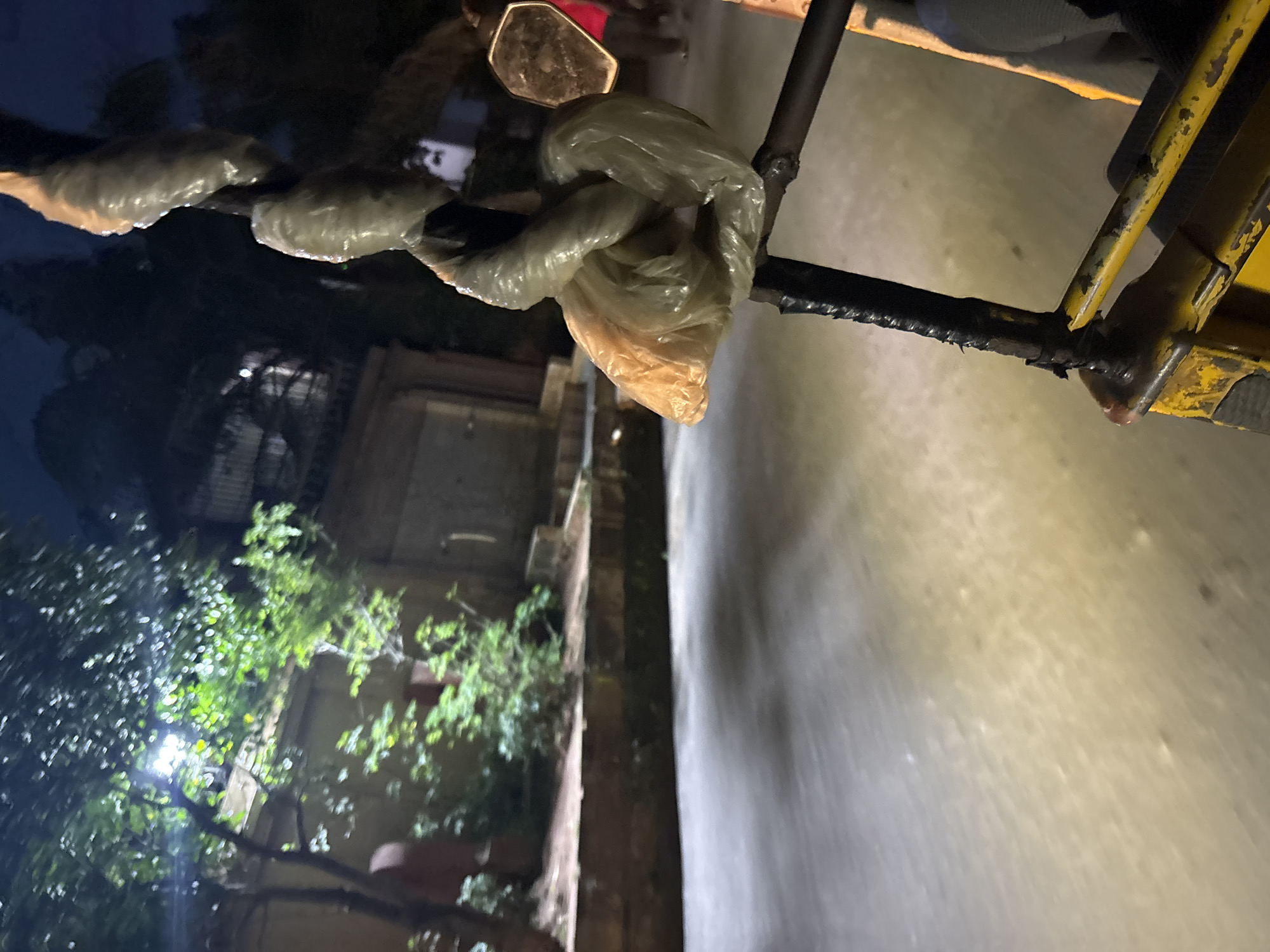 The image depicts a nighttime scene in Onitsha, Nigeria, likely from the perspective of a passenger in a vehicle. In the foreground, a metal bar is wrapped with black tape and then encased in plastic bags, one of which is translucent orange and appears to be tied around the bar. The road surface in front of the vehicle is wet and reflects the available light. To the left, trees with green foliage are illuminated by a bright light source, casting shadows. Behind the trees, a brick structure with a closed doorway is visible. In the background, darker shapes suggest buildings and trees against a night sky. A rear-view mirror is partially visible on the right side of the frame, reflecting a person in a red shirt. Another person is partially visible in the lower right corner, seated within the yellow vehicle. The overall impression is of movement on a wet road at night, with urban and natural elements present.