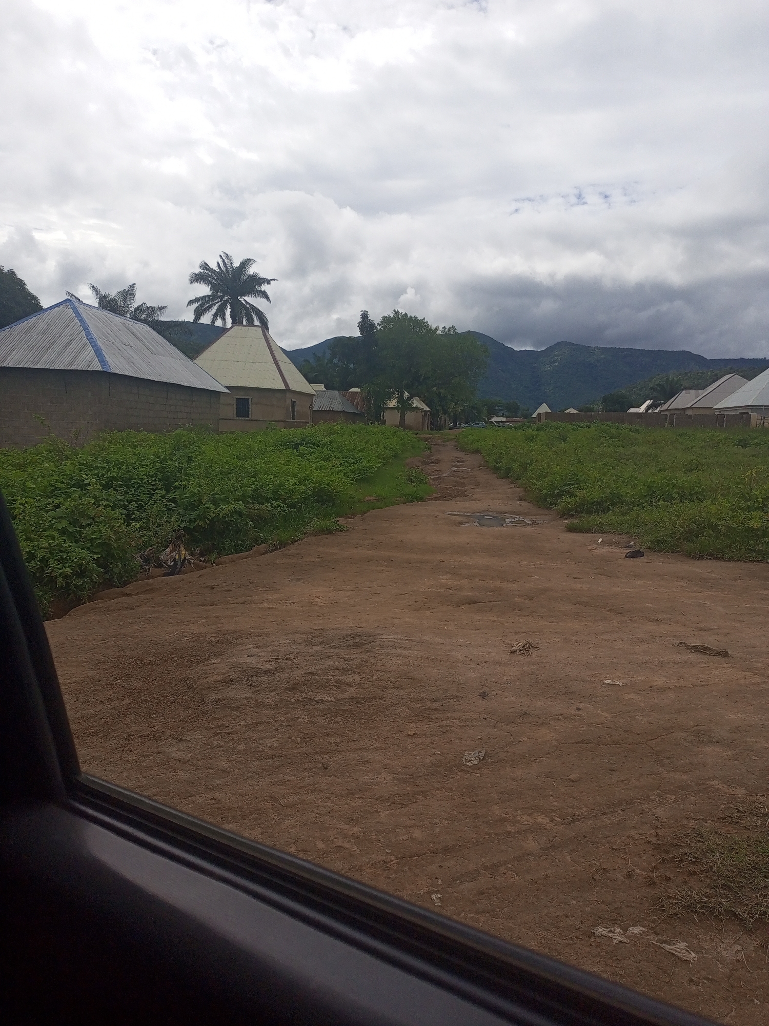 The image depicts a dirt road leading through a village in Serti, Nigeria. The road is flanked by dense green vegetation on both sides. Several buildings with corrugated metal roofs are visible in the distance. One building to the left has a conical, light-colored roof. A palm tree stands prominently near the center of the image, with other trees and hills forming the backdrop. The sky is overcast with heavy clouds. The perspective suggests the photograph was taken from inside a vehicle, with the edge of a car door visible in the foreground. Scattered debris, appearing to be plastic bags and other refuse, is present along the roadside and on the dirt path. The overall environment appears rural and somewhat undeveloped.