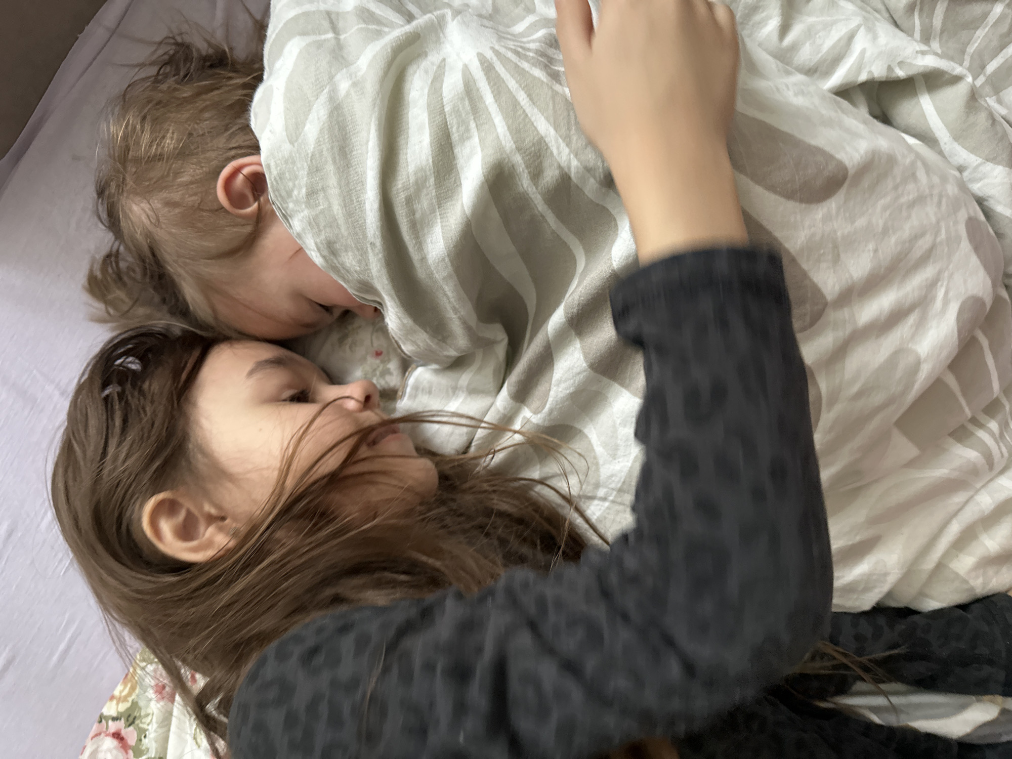 This intimate overhead shot captures two children sleeping or resting together in a bed. The scene is peaceful and quiet, likely set indoors in a bedroom.
The older child, positioned in the foreground and to the left, has long, dark brown hair that partially covers their face. They are lying on their right side, with eyes closed or nearly closed, and their face suggests deep rest. They wear a dark, long-sleeved top with a subtle, possibly leopard or animal print pattern. Their right arm is raised, with the hand partially visible and resting near their head or under a blanket.
The younger child is nestled close to the older one, positioned slightly above and to the right, almost completely covered by a light-colored duvet or blanket. Only the top of their head, a ear, and a small part of their face are visible. They have lighter, possibly blond or light brown, hair. They appear to be sleeping soundly, their face turned towards the older child.
The bedding consists of several layers: a plain light-colored sheet on the bed, a floral-patterned fabric (possibly a pillowcase or bottom sheet) under the older child's head, and a white and grey/beige striped duvet or blanket covering both children, particularly the younger one. The overall impression is one of comfort and closeness.
No visible text is present in the image. The light suggests it could be morning or evening, with soft, natural lighting.