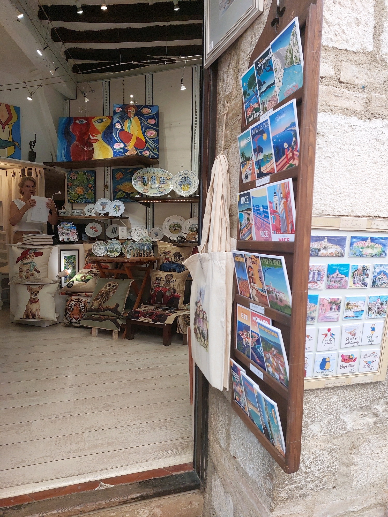 This is a medium shot of an outdoor art and souvenir shop in Saint-Paul-de-Vence, France, with a focus on the display of postcards and art. The shop is open to a stone street, with a wooden floor inside. A woman with blonde hair, wearing a white top, stands behind a counter to the left, holding a piece of paper. In the background, colorful paintings are displayed on the wall, including a large abstract piece with a woman's face. Shelves are laden with decorative plates and cushions. To the right, a wooden rack displays a variety of vintage-style postcards featuring scenes from various French locations. A framed collection of small paintings or prints is mounted on the stone wall next to the postcard display. The overall impression is one of a charming, traditional art boutique.
