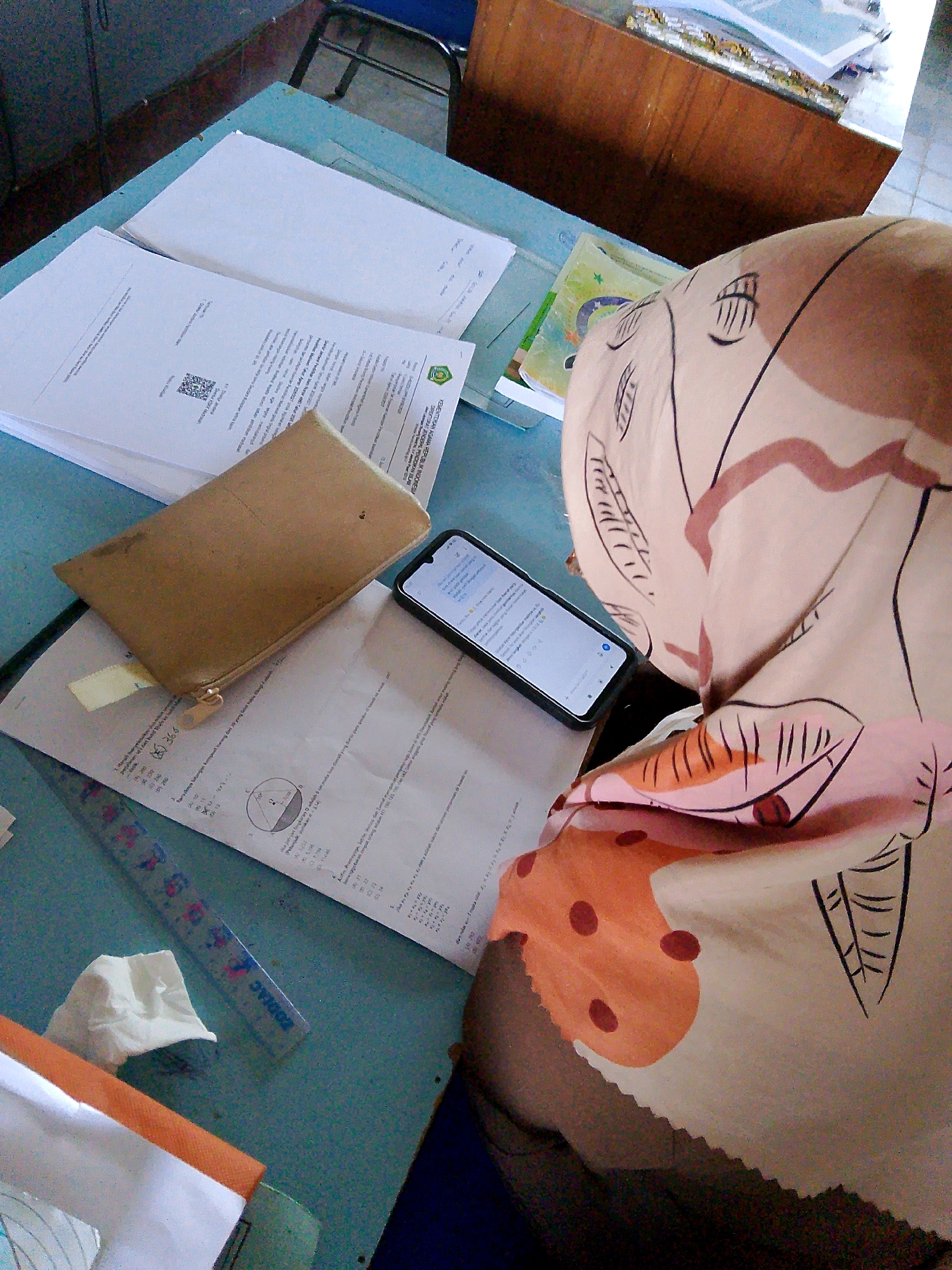 The image depicts a close-up, overhead view of a blue desk in what appears to be a classroom or study area. The desk is covered with various documents, including papers with text and diagrams, a brown zippered pouch, and a smartphone lying face up, displaying a screen with text. A ruler with measurement markings is also visible on the desk. In the background, a wooden cabinet is present. A portion of a person's arm, covered by a light-colored fabric with a pattern of abstract shapes and lines in brown and pink, is visible in the foreground. The overall scene suggests a workspace or a moment of study. There is no discernible information within the image that indicates the specific location of Kuripan Utara, Indonesia.