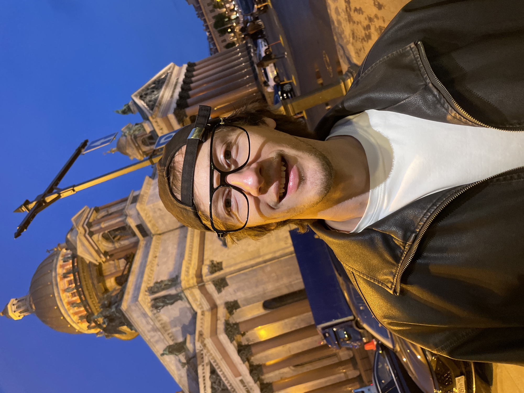 A young man, wearing a black baseball cap, glasses, a white t-shirt, and a black leather jacket, takes a selfie in front of St. Isaac's Cathedral in Saint Petersburg, Russia. The sky is dark blue, indicating it is either dusk or night. The cathedral, a large neoclassical building with a prominent golden dome, is illuminated by warm lights. In the background, cars and a blue truck are visible on a street, and streetlights cast a yellowish glow on the scene. The man is smiling at the camera.