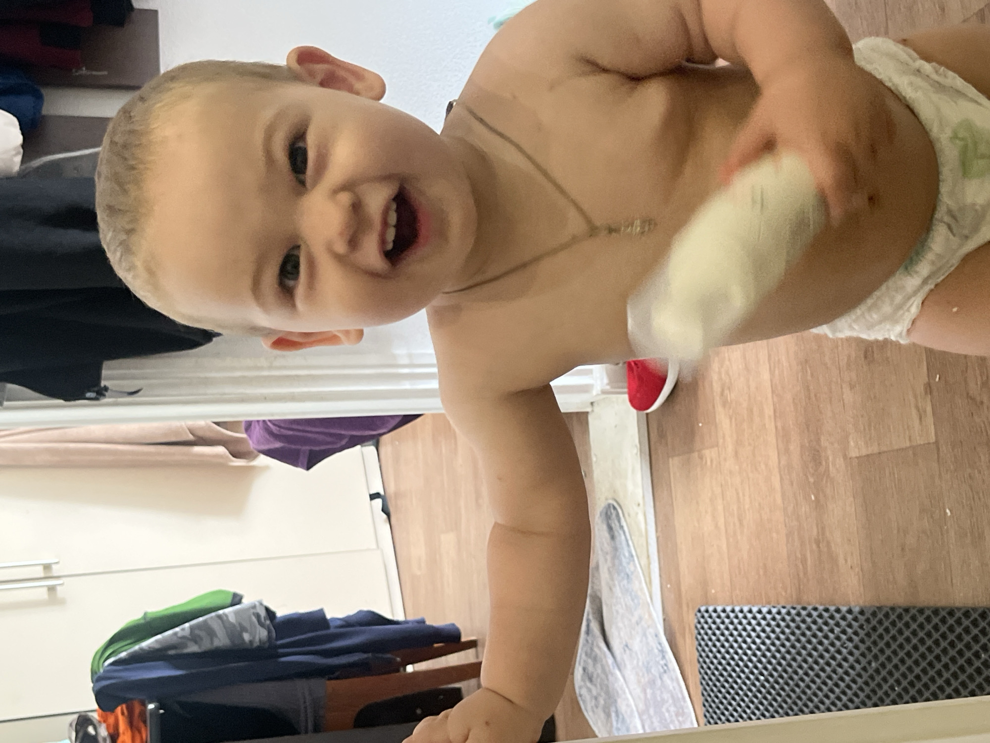 The media file shows a close-up of a baby in a diaper, standing upright. The baby has fair skin, blonde hair, and is looking directly at the camera with a wide smile, showing their teeth. They are holding a white cylindrical object in their right hand, which appears to be a baby wipe dispenser or similar product. A thin, light-colored necklace is visible around the baby's neck. The baby is positioned in a room with wooden flooring. In the background, there is a closet with doors and shelves containing clothes, including a purple shirt, green and grey patterned fabric, and navy blue garments. A red shoe is visible near the baby's right foot. The overall scene suggests a domestic setting, likely a bedroom or dressing area, with natural lighting. The baby's expression conveys joy and playfulness. There is no visible text in the image. The location context is Chelyabinsk, Russia.