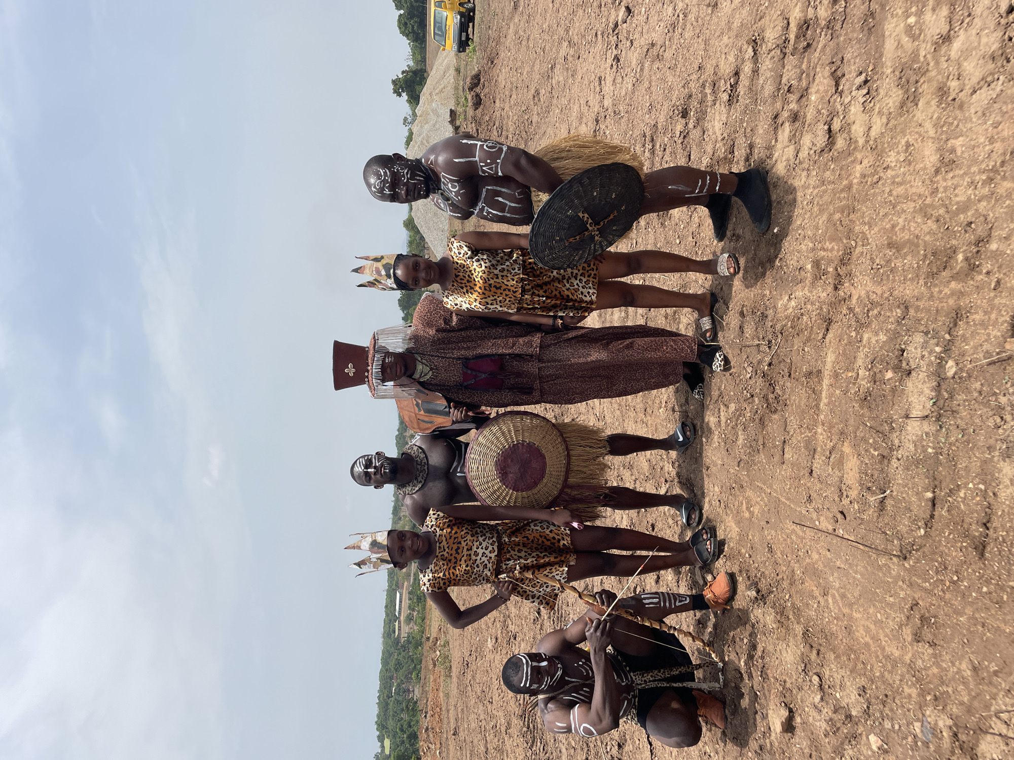 A photo taken in Akwanga, Nigeria, shows six individuals in traditional attire posing outdoors. The ground appears to be dirt and earth.
A man at the top of the frame and another at the bottom are kneeling, both with white markings on their faces and bodies. The top kneeling man has a black shield in his hand. The kneeling man at the bottom has a bow in his hand and is in the act of aiming or shooting an arrow.
Two women, wearing matching leopard print dresses and headpieces, stand between the kneeling men.
One taller man stands in the middle, wearing a long, leopard print robe and a ceremonial hat.
Another tall man stands next to one of the women, holding a large, round wicker shield.
A yellow vehicle is visible in the background.