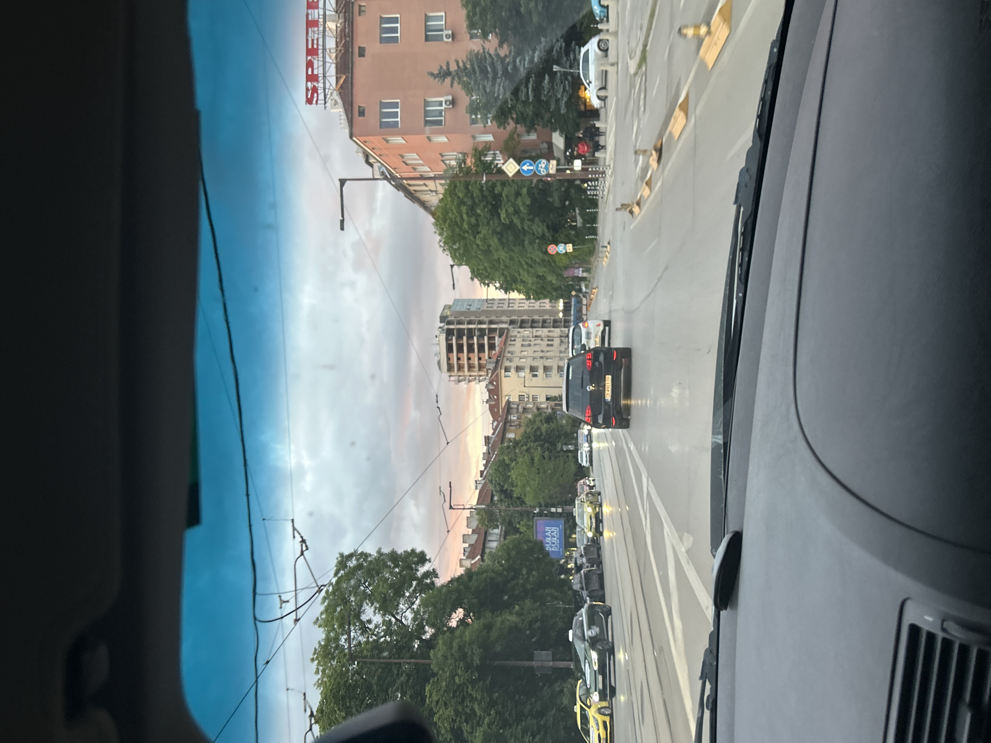 The image, taken from inside a vehicle looking forward, captures a street scene in an urban environment. The sky above is cloudy with hints of pink and orange, suggesting either dawn or dusk. Overhead, power lines and tram wires crisscross the sky.
In the foreground, the dashboard of the vehicle is visible, with a dark object, possibly a phone mount or a small electronic device, positioned on it. The windshield shows some smudges.
The street is paved and appears wet, reflecting the surrounding lights and sky. Multiple cars are on the road, including a black SUV in the center lane, a yellow taxi on the left, and other vehicles of various colors. Some cars have their headlights on. Tram tracks are visible on the left side of the road.
Buildings line both sides of the street. On the right, a multi-story pink building with visible windows and air conditioning units stands tall, topped with a red sign that reads "SPETER". Further down the street on the left, a partially constructed building with exposed framework is visible. Trees and greenery are interspersed among the buildings and along the roadside.
Various traffic signs and cones are present, indicating road markings, traffic flow direction, and potential construction or lane closures. Pedestrians are visible on the sidewalk in the middle distance on the right. The overall impression is a busy city street during transitional lighting conditions.