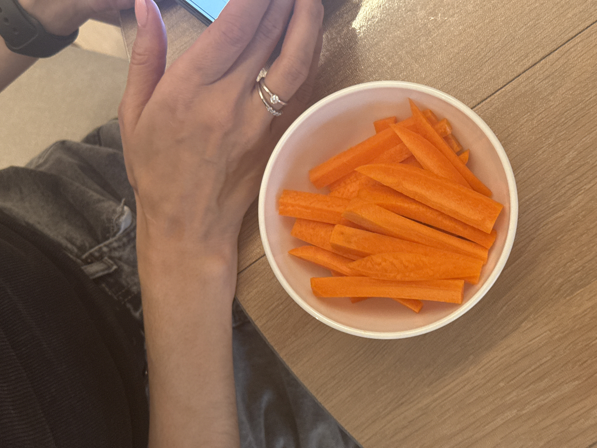 An indoor scene captured from an overhead perspective shows a person's hands and lower body next to a bowl of carrots on a wooden table. The person, likely in Perm, Russia, is holding a smartphone with their left hand, which has light pink painted fingernails. On the ring finger of this hand, two rings are worn: a silver-colored band with a small clear stone, and a plain band stacked beneath it. A black smartwatch or fitness tracker is visible on the person's left wrist. The individual is wearing dark clothing, possibly jeans or casual trousers.
To the right of the person's hands is a light-colored, possibly white or pale pink, bowl filled with numerous orange carrot sticks or julienned carrots. The bowl rests on a light brown wooden table with visible grain patterns. The overall activity suggests a relaxed moment, possibly snacking while using a mobile device. No specific time of day, weather conditions, or visible text can be determined from the image. Emotions are not discernible.