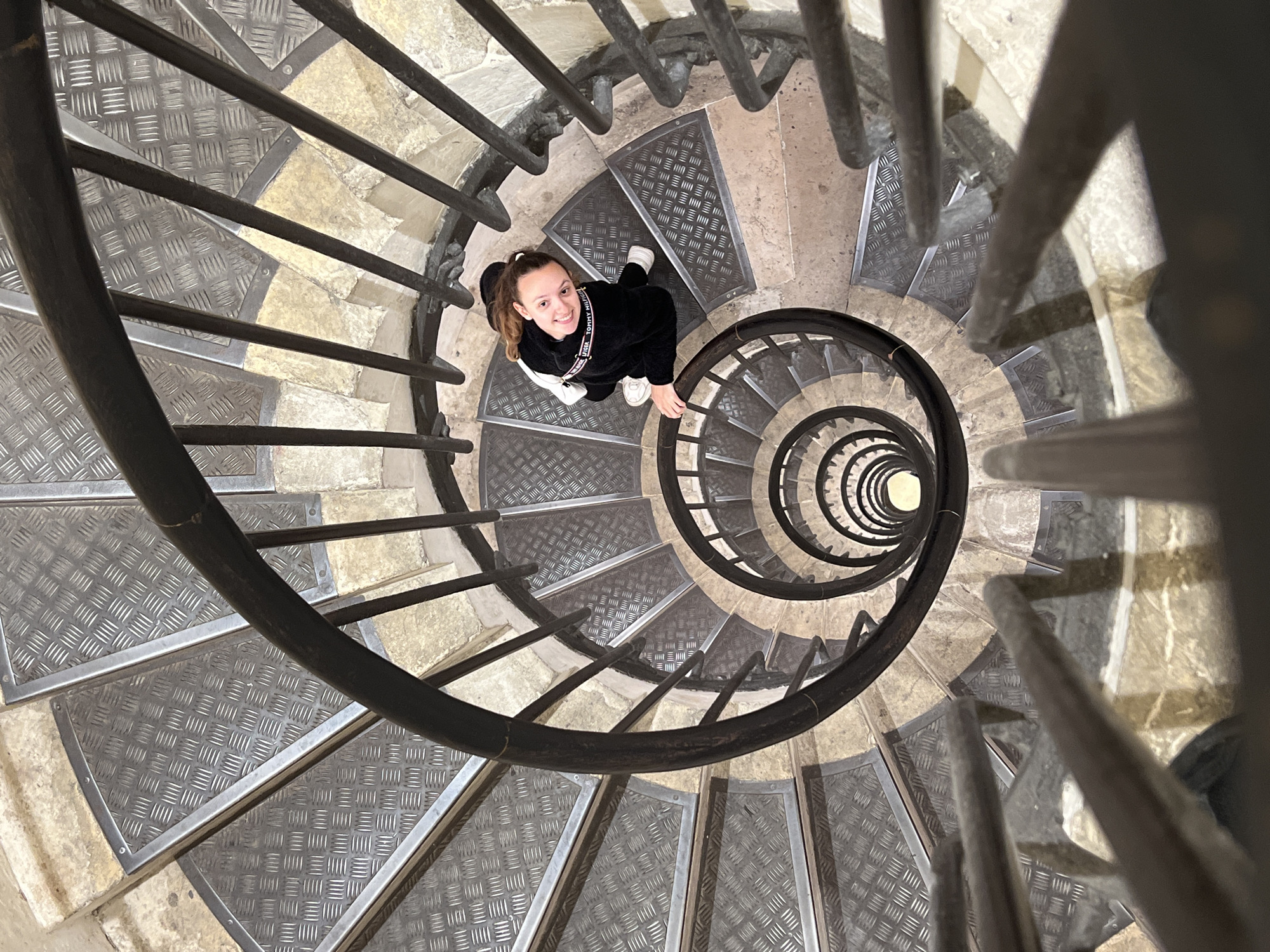 An overhead, worm's-eye view captures a young woman standing on a spiral staircase, looking up and smiling brightly towards the camera. She has light brown hair tied up and is wearing a black long-sleeved top, white sneakers, and a white bag strap across her body. The brand "TOMMY HILFIGER" is visible on the neckline of her top.
The staircase is a prominent feature, coiling downwards into a tight, dizzying spiral, creating a strong sense of depth. The steps are made of light-colored stone with dark grey metallic anti-slip plates (chequer plate pattern) embedded in them. A sturdy, dark metal handrail follows the curve of the staircase, reinforcing the spiral effect. The scene suggests an interior location, likely a historic building or tower, consistent with a location in Paris, France. The upper sections of the staircase are well-lit, with light gradually diminishing as the spiral descends. The image combines human presence with striking architectural detail.