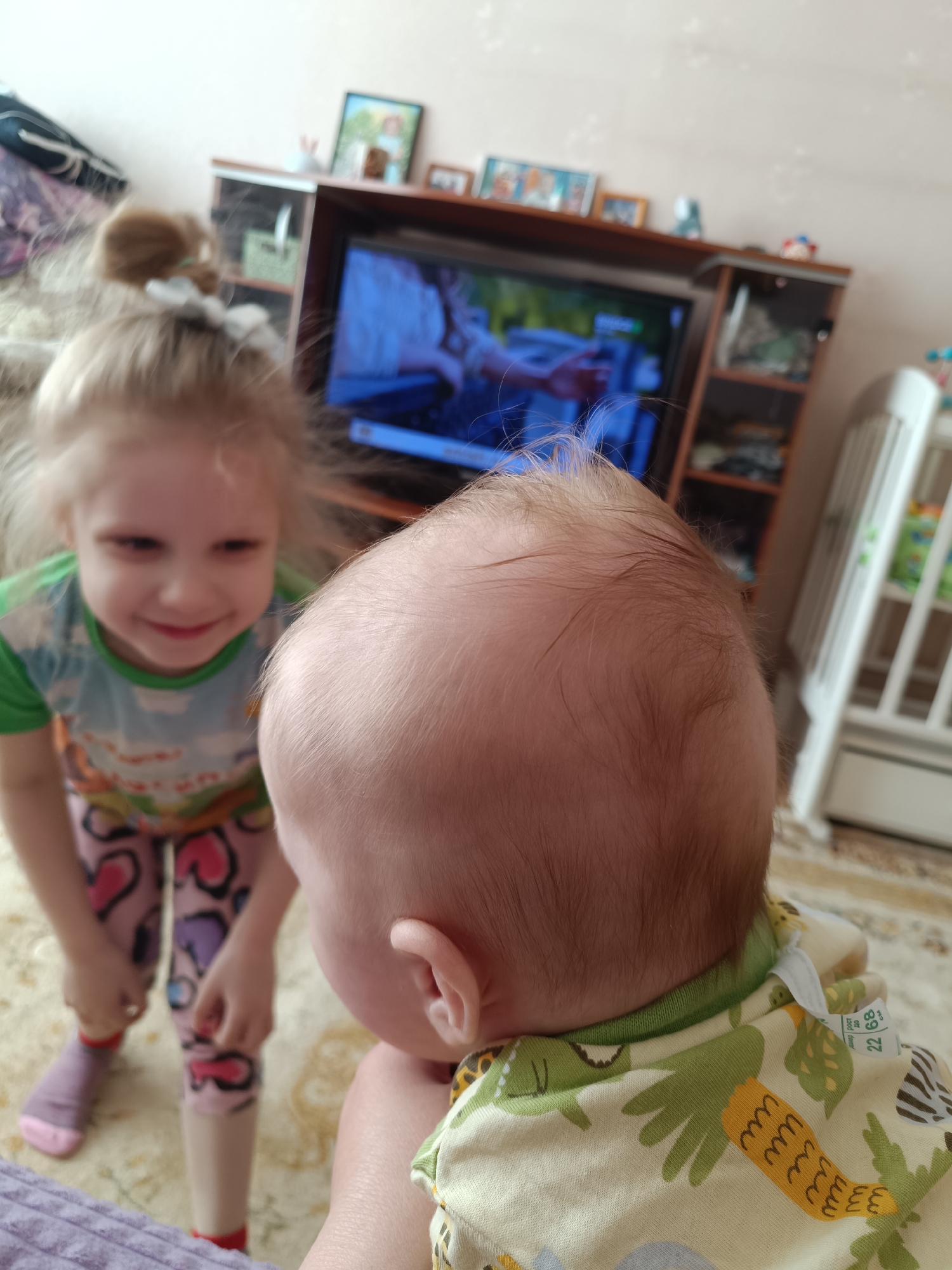 A photograph shows a young, fair-skinned child with blonde hair pulled into a ponytail, wearing a green and multicolored shirt and pink leggings, smiling and looking down at a baby. The baby, with a fair, bald head and light brown fuzz of hair, is facing away from the camera. The baby is wearing a light-colored outfit patterned with green and brown.
In the background, there is a carpeted floor, a television on a stand with photographs and objects on top of it, and a white crib.
There is no evidence to determine this photograph was taken in Tobolsk, Russia.