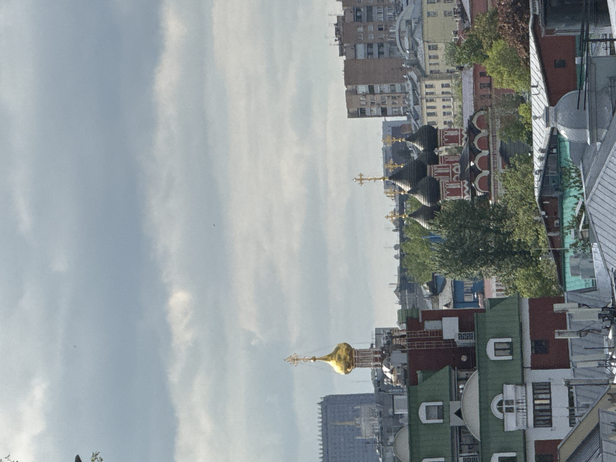 The image shows a cityscape in Moscow, Russia. The focal points are the domes and spires of Russian Orthodox churches, with distinct architectural features, including black onion domes with gold crosses, and a separate gold dome. A range of buildings of varying heights and colors are present, including residential and commercial structures. The sky is overcast with a layer of clouds.