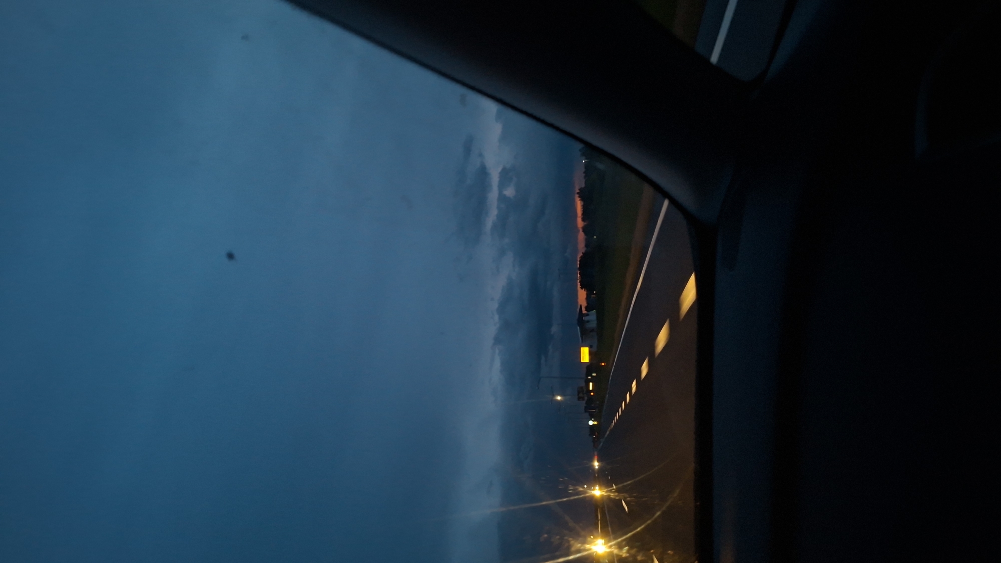 The image depicts a view from inside a vehicle, likely a car, at dusk or twilight. A road stretches into the distance, illuminated by streetlights. The sky is a mix of dark blue and grey clouds, hinting at a recent or impending rain. The location is possibly within or near Дабрынёўскі сельскі Савет, Belarus, given the presence of road infrastructure and ambient lighting characteristic of a rural or semi-urban area. There are no visible people or specific interactions within the frame. The scene appears to be a calm moment during transit.