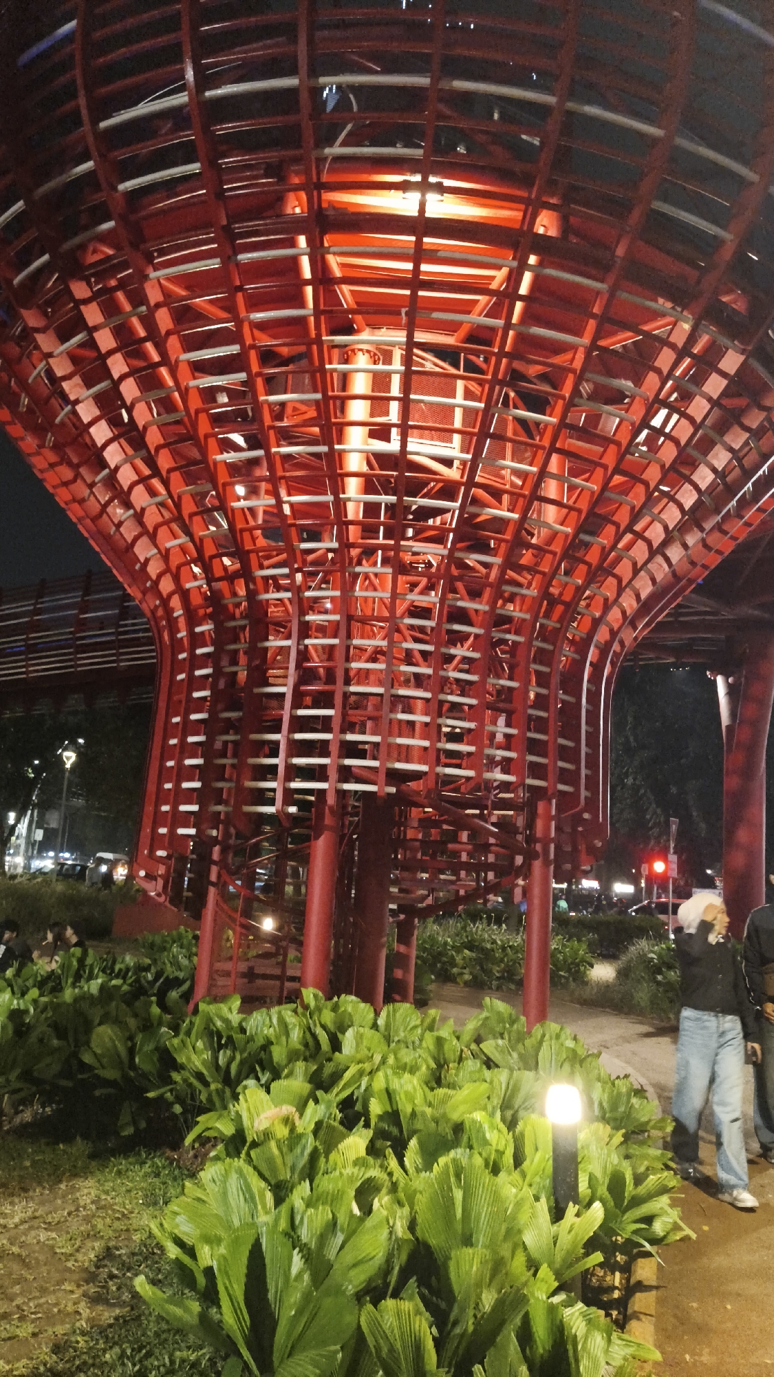 A red, abstract architectural structure with a curved, cage-like design dominates the frame, illuminated from within with orange light. The structure appears to be a pedestrian bridge or walkway, with spiraling stairs visible inside its base. Lush green foliage, resembling large palm leaves, carpets the foreground. In the background, people are visible walking and standing near the structure. The scene is at night, with streetlights and traffic signals indicating an urban environment. The location is identified as Jembatan Penyeberangan Orang (JPO) Istora, Gelora Bung Karno, South Jakarta, Indonesia.