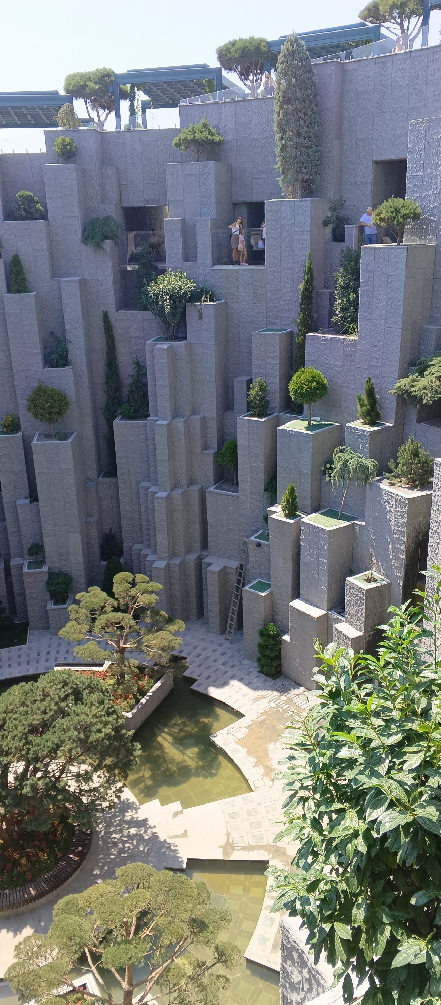 A high-angle, outdoor shot captures a modern architectural design featuring stepped, stone-clad structures with numerous recessed openings and planters. These structures create a vertical, canyon-like effect, with various trees and shrubs strategically placed within planters at different levels. In the foreground, a large tree with dense green foliage casts a shadow on a paved area surrounding shallow, greenish pools of water.
Several people are visible within the recessed openings of the stone structures. On a higher level, a couple stands near a railing, observing the scene. Further down, a group of three individuals, including two adults and a child, are gathered in an opening. Another adult is visible in a recessed area on the right.
A metal ladder leans against one of the stone structures, suggesting access to higher levels. The paving stones are light-colored and arranged in a patterned walkway around the water features. The overall impression is of a meticulously designed public space that integrates architecture and horticulture, possibly a garden or a museum complex. The sky is clear and bright, indicating a sunny day.