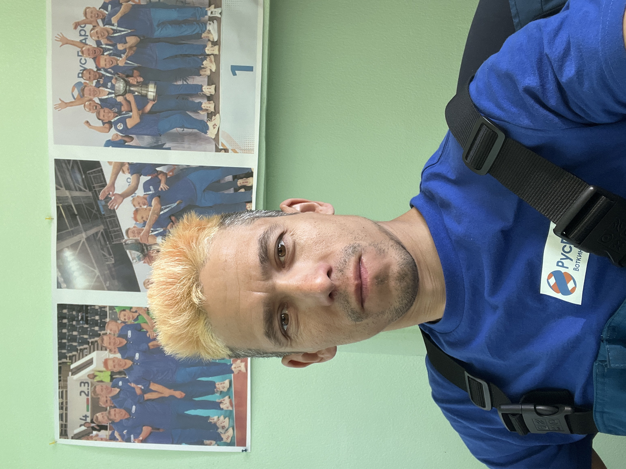 The image shows a man with dyed blonde hair and a blue t-shirt standing indoors in front of a light green wall. He has brown eyes and stubble. The t-shirt has a logo with the word "Рускин" above the word "Воткин" on it. Straps are visible over his shoulders. On the wall behind him are three photographs of a group of people wearing blue uniforms. The top photo is labelled with a "1". The bottom photo is labelled with "4-2.3". The photograph was most likely taken in Chaikovsky, Russia, due to the Russian writing on the t-shirt logo.