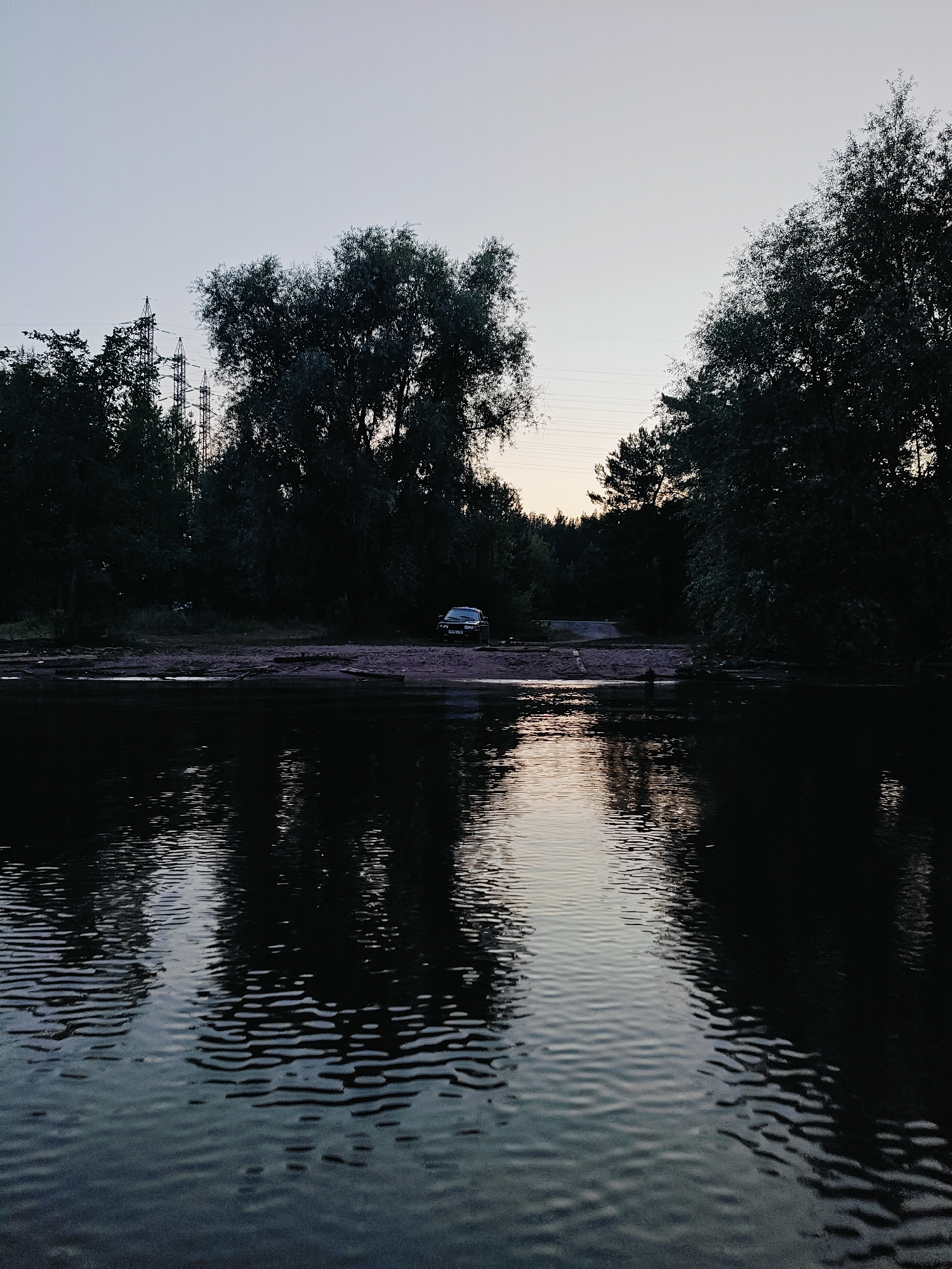 The image depicts a serene riverside scene at dusk in Noviy, Russia. The foreground is dominated by dark, rippling water, reflecting the fading light of the sky and the silhouettes of trees. Beyond the water, a sandy bank leads to a dense forest. A single, dark-colored car is parked on the bank amidst the trees, suggesting a moment of quiet respite or an arrival.
The sky is a pale, soft gradient of blue and grey, indicative of twilight. Tall power line towers are visible in the distance, breaking the natural skyline and hinting at the proximity of civilization. The overall mood is peaceful and contemplative, with the stillness of the water and the soft light creating a tranquil atmosphere. The lack of human presence, aside from the parked car, adds to the sense of solitude. There is no visible text in the image.