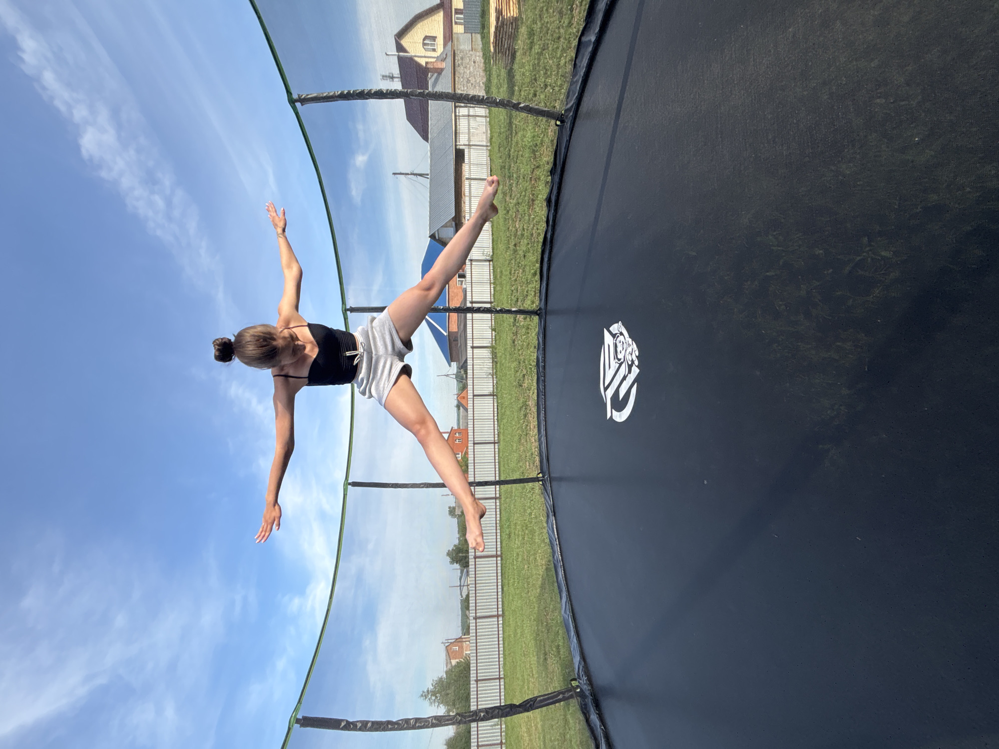 A young woman is captured mid-air, jumping on a black trampoline with a safety net enclosure. She is wearing a black tank top and light grey shorts, with her arms extended horizontally and her legs spread apart in a straddle split. Her hair is styled in a bun. The trampoline is situated outdoors on a grassy area, with a white picket fence and houses visible in the background. The sky above is blue with scattered white clouds. The scene appears to be in a rural setting, possibly the city of Vishnevka, Russia, given the architecture of the houses.