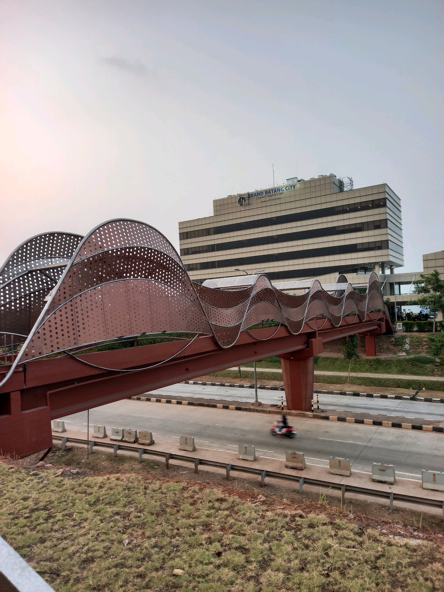 The image shows an elevated pedestrian bridge with a distinctive wavy, perforated metal design in shades of reddish-brown and gray. The bridge spans over a road where a single motorcyclist is visible, blurred by motion. In the background, a large, multi-story building with a light-colored facade and dark horizontal window bands is prominent. The building has signage that reads "GRAND BATANG CITY" in blue lettering. The scene appears to be in an urban environment, likely Ketanggan, Indonesia, given the context provided. The sky is overcast with a pale, diffused light. The foreground consists of a grassy embankment.