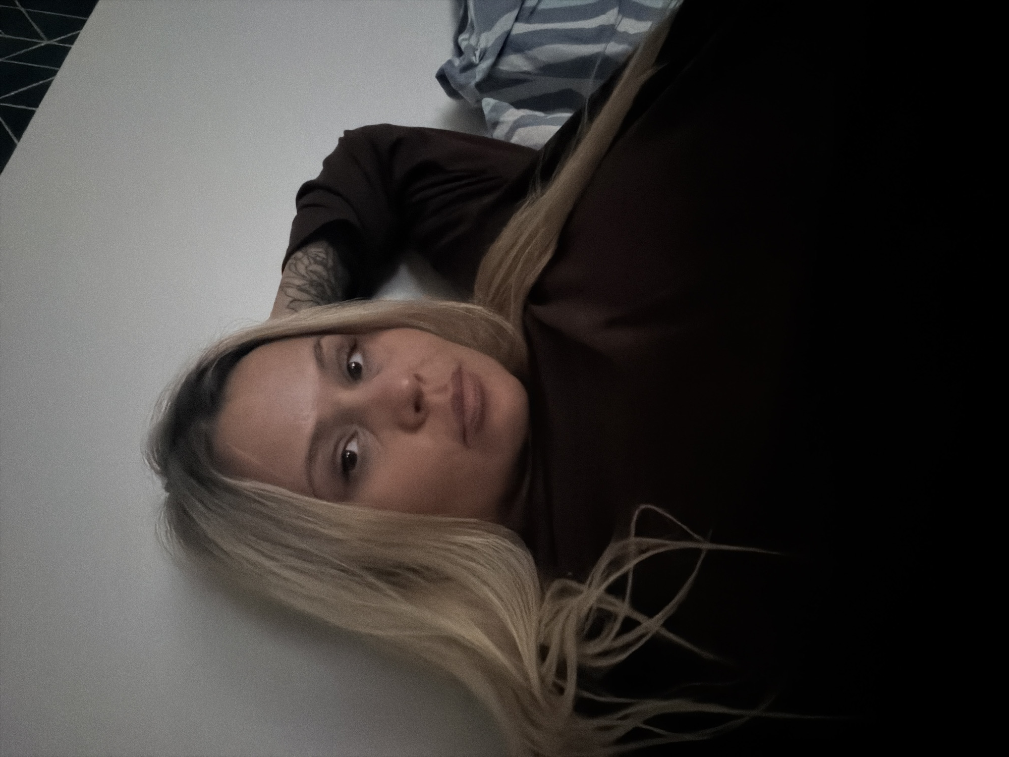 A woman with long, blonde hair lies on a white surface. She is wearing a dark brown top. Her left arm, visible, has a tattoo. A striped fabric is visible in the background. The location is unknown and there is no indication that it is the city of Sarapul, Russia.
