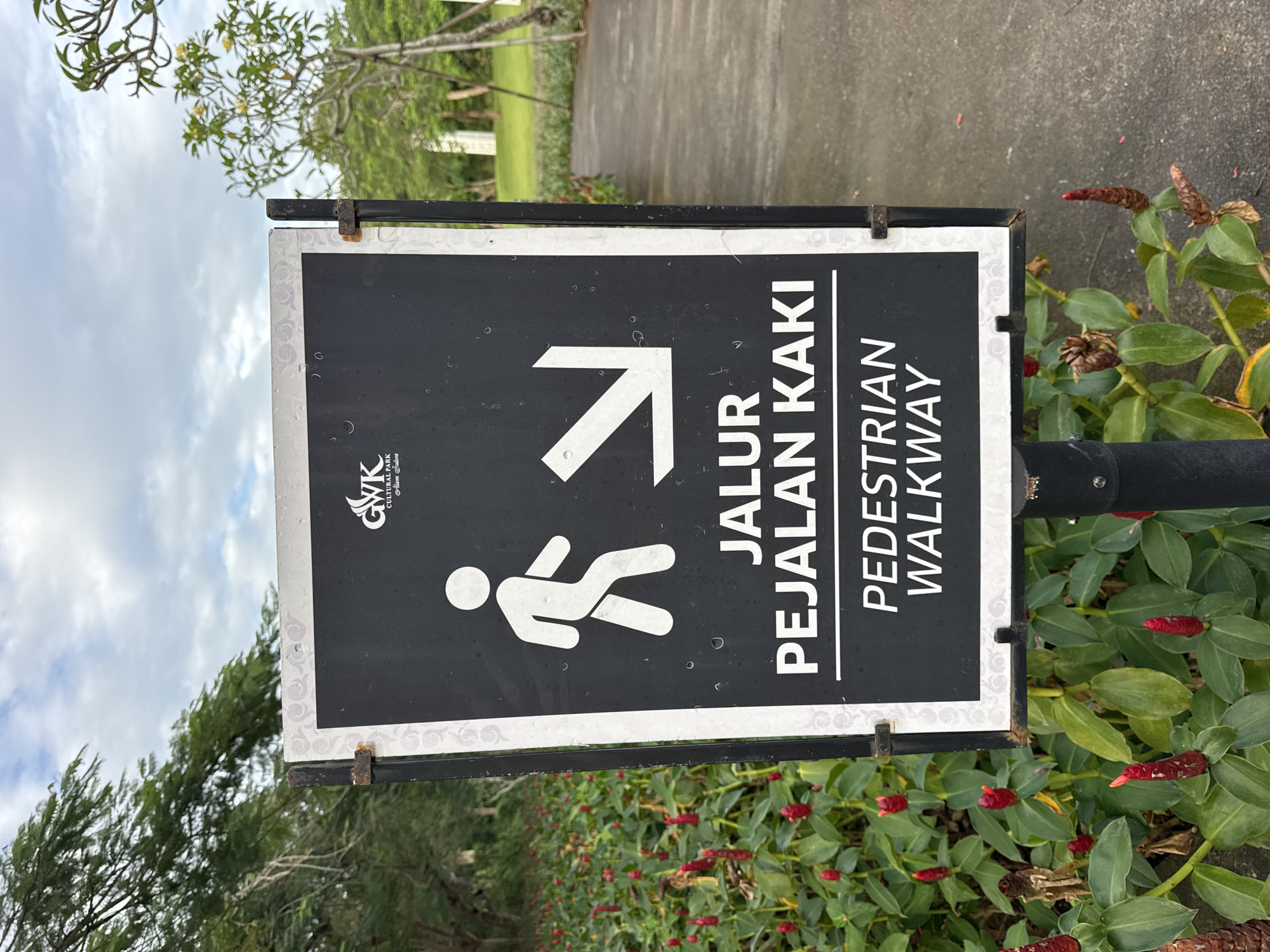 A close-up, slightly angled shot shows a sign in Kuta Selatan, Indonesia, indicating a pedestrian walkway. The sign is rectangular, with a black background and white text and symbols. A white pedestrian figure and an arrow pointing diagonally upwards and to the right are on the left side of the sign. To the right of these, in large, bold white text, are the words "JALUR PEJALAN KAKI" and "PEDESTRIAN WALKWAY." The sign also features a logo, "GWK CULTURAL PARK - Uluwatu Bali," in a smaller font above the pedestrian figure.
The sign is mounted on a dark metal pole. Green foliage and red flowers surround the base of the pole. In the background, a paved walkway leads into a blurred landscape of trees and grass. The sky is visible, with scattered clouds. The sign appears to be slightly weathered.