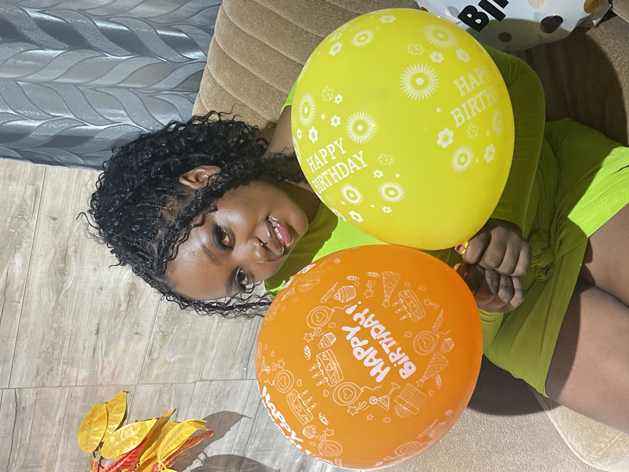 The image depicts a joyous birthday celebration featuring a person with dark skin and dark, braided hair, lying down and smiling directly at the camera. They are wearing a bright, lime-green garment. A nose piercing is visible on their face.
Two prominent "Happy Birthday" balloons are in the foreground. The larger one is yellow, decorated with white floral and star patterns, and features the text "HAPPY BIRTHDAY" in white. Below it is an orange balloon, covered with white outlines of birthday cakes, party hats, gifts, and stars, also bearing the inscription "HAPPY BIRTHDAY!". A third white balloon, partially visible in the top right corner, also displays part of the word "BIRTHDAY" and polka dot patterns.
The scene appears to be an indoor, domestic setting. The individual is resting on a beige, ribbed sofa or couch. The flooring consists of light-colored wooden planks. In the lower left, a decorative plant with vibrant yellow and red leaves adds a touch of color. A grey patterned wall or curtain is visible in the background. The lighting suggests an indoor environment, possibly under artificial light, creating soft shadows. The overall mood conveyed is celebratory and happy.