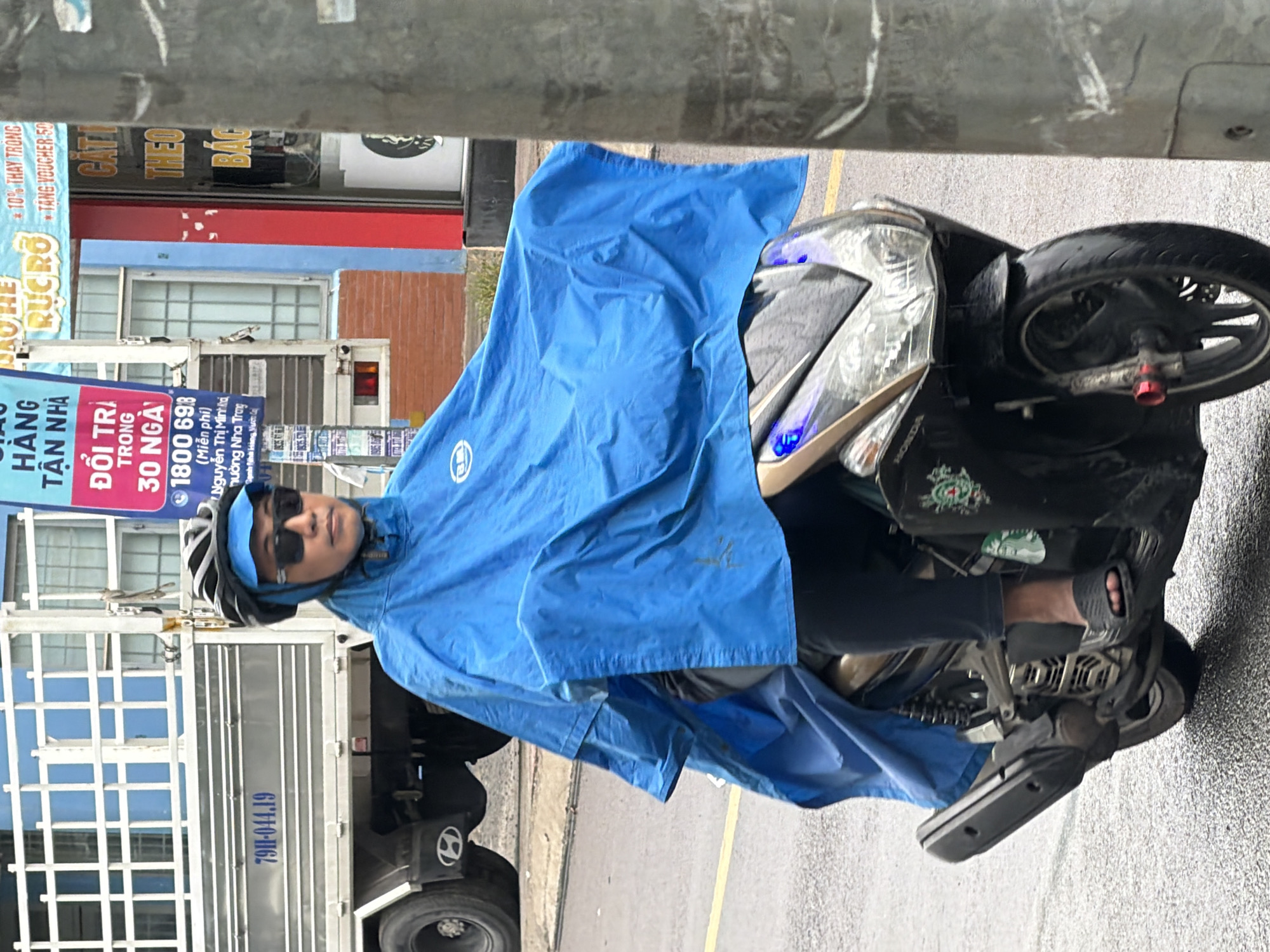 A person wearing a blue raincoat and helmet rides a black motorcycle on a wet street in Nha Trang, Vietnam. The motorcycle has a "Honda" logo and blue lights on the front. The person is wearing sunglasses and black pants. A large white truck with the license plate "79H-044.19" is parked behind the motorcycle. A red and blue sign with Vietnamese text is visible on the side of a building in the background. A metal pole stands to the right of the image. The street is wet, indicating it is raining or has recently rained.
