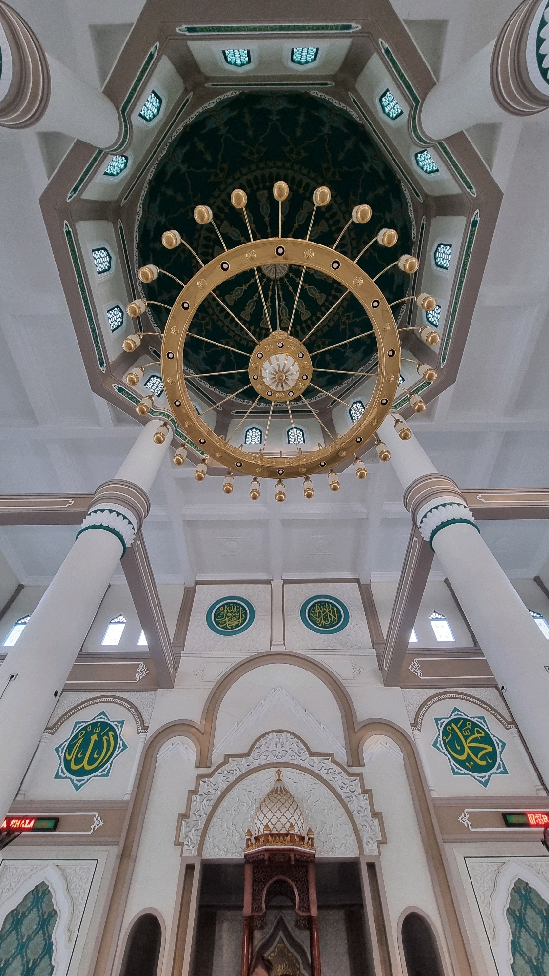 The image shows an interior view of the Islamic Center of Lhokseumawe, located in Lhokseumawe, Indonesia. The perspective is from below, looking upwards towards the ceiling and a large, ornate chandelier. The chandelier is circular and gold-colored, with many lights. The ceiling is intricately decorated with green and gold patterns and features several small, square windows. Two massive, white columns with decorative bands flank the central area, supporting the ceiling structure.
Below the chandelier, the architecture features arches and decorative Islamic calligraphy inscribed on the walls. In the foreground, on the lower part of the image, a decorative structure resembling a minaret or a decorative pulpit with a golden dome is visible. There are also digital displays indicating time. The walls are painted in light colors with brown accents, and some areas feature geometric patterns and floral motifs. The overall impression is one of grandeur and religious significance within the mosque. No people are visible in the image.