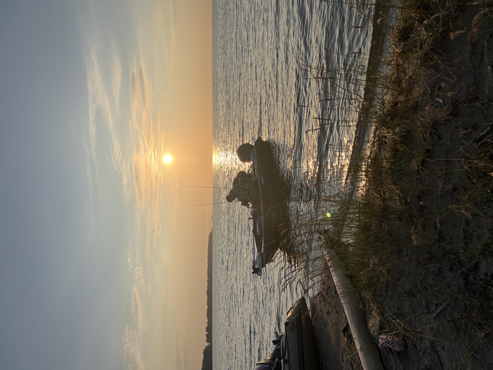 A fishing boat is docked on the shore of a lake at sunset. The sun is setting on the horizon, casting a warm glow over the water. The boat is a small, aluminum vessel with a trolling motor. A person is seated in the boat, wearing camouflage clothing and holding a fishing rod. Another, smaller boat is partially visible on the left side of the image, also docked on the shore. The shore is sandy and covered with sparse vegetation. The scene appears to be in a rural area, with a treeline visible in the distance across the lake. The location is specified as Borovoe, Russia.