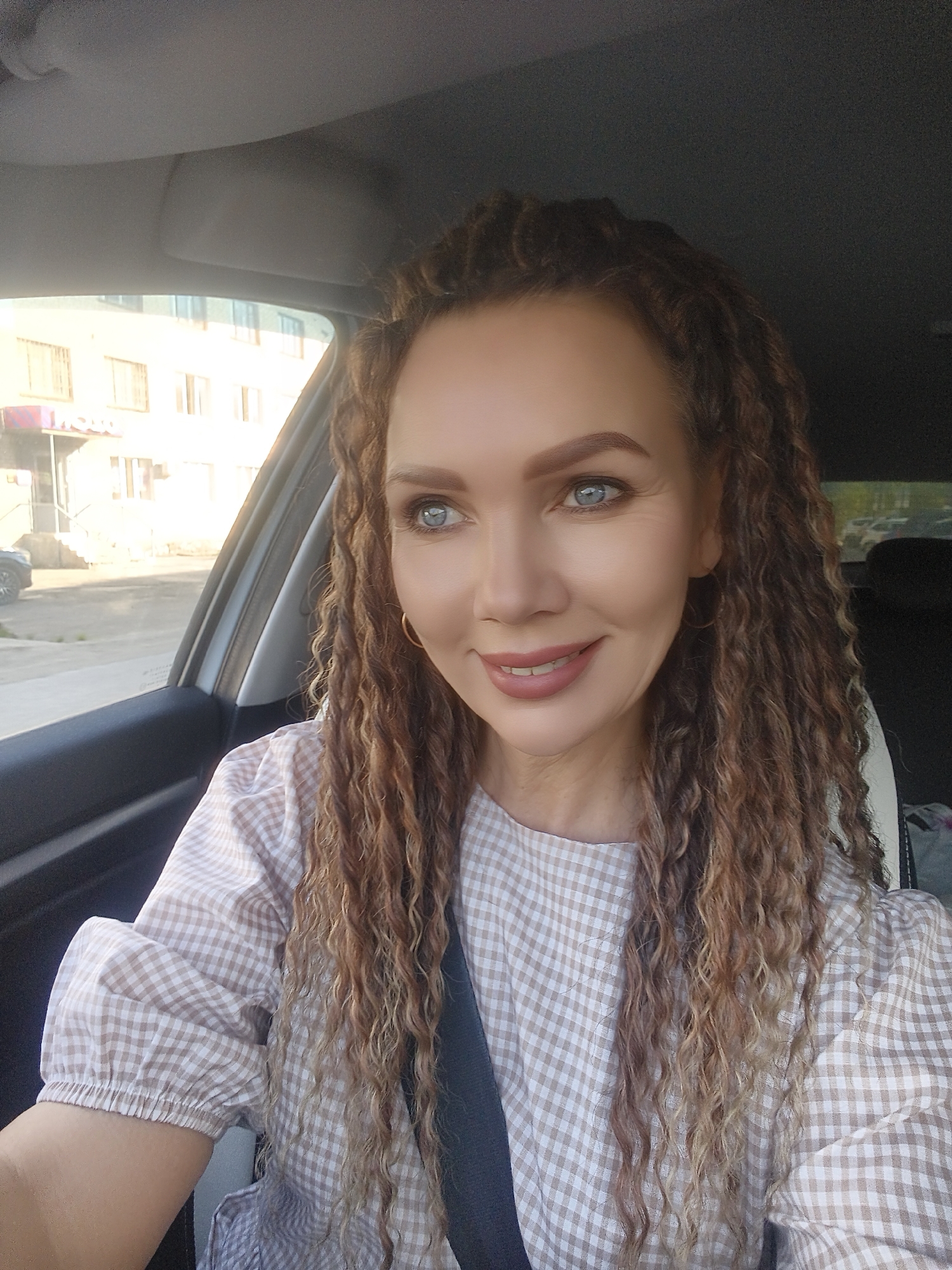 A light-skinned woman with light blue eyes and curly brown and blonde hair is seated in the driver's seat of a car, wearing a tan and white checkered top and a black seatbelt. She is looking to the right of the frame with a slight smile. Outside the car window, there is a building with multiple windows and a car parked nearby. The location appears to be an urban setting, within the city of Unknown, Unknown.