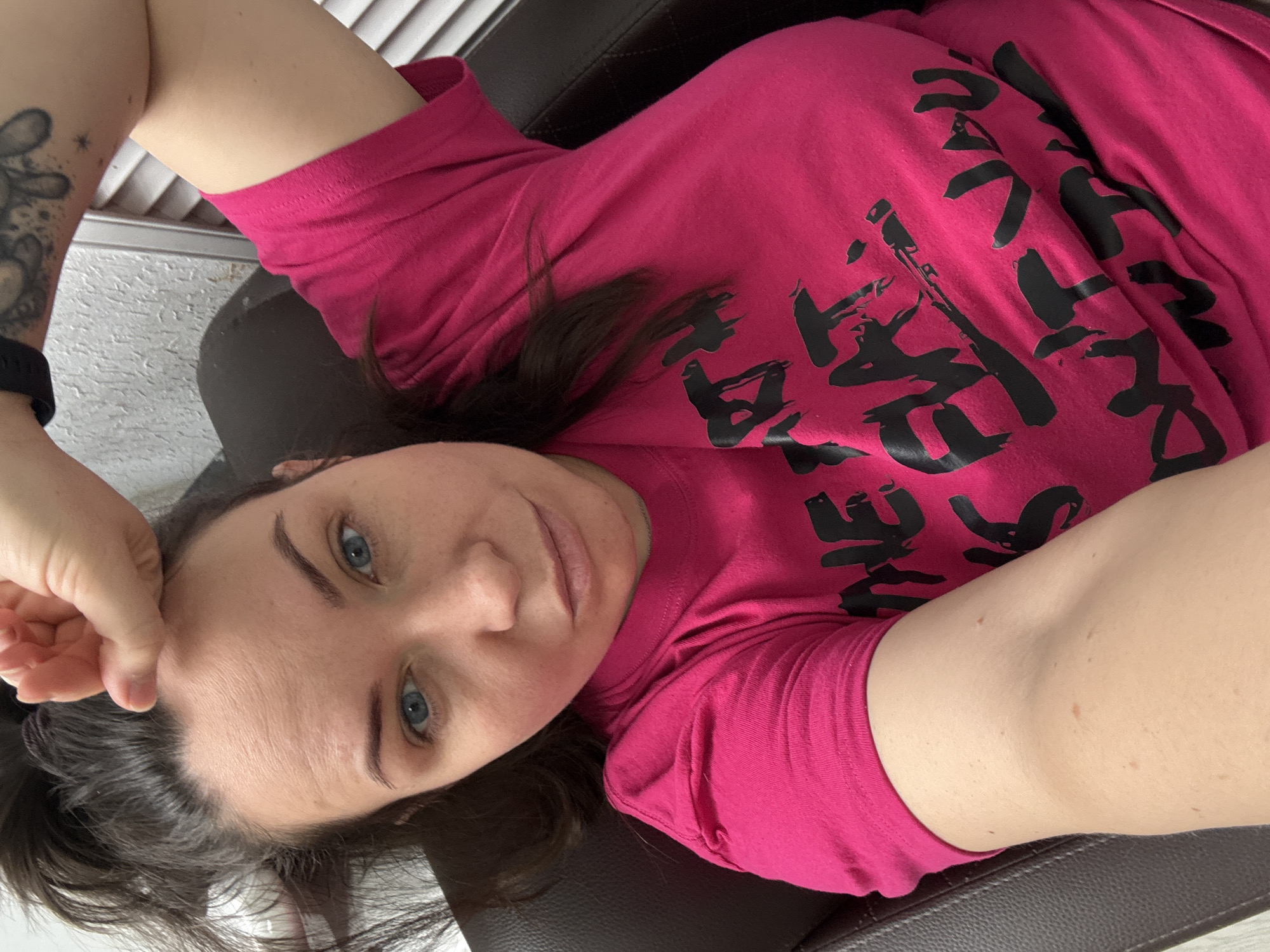 A woman is featured in a high-angle, close-up selfie, lying down on a dark brown or black surface, likely a chair or sofa. She wears a bright pink t-shirt adorned with stylized black text on the front. Her head is tilted, and she gazes directly at the camera with a neutral expression, showcasing her light blue eyes. Dark hair frames her face and rests on her shoulders and shirt.
Her left forearm reveals a tattoo featuring a cartoonish figure, possibly a rabbit, along with small star designs. A dark wristband, resembling a watch or fitness tracker, is also visible on her left wrist. The background is softly blurred but shows white horizontal blinds or louvers and a textured, light-colored wall or surface, indicating an indoor setting.
The black text on her t-shirt is rendered in a brush-stroke style. While challenging to fully discern, some characters appear to be Japanese Katakana, potentially spelling "ブラック" (burakku), meaning "black." The image conveys a casual, relaxed atmosphere, suggesting a self-portrait taken indoors.