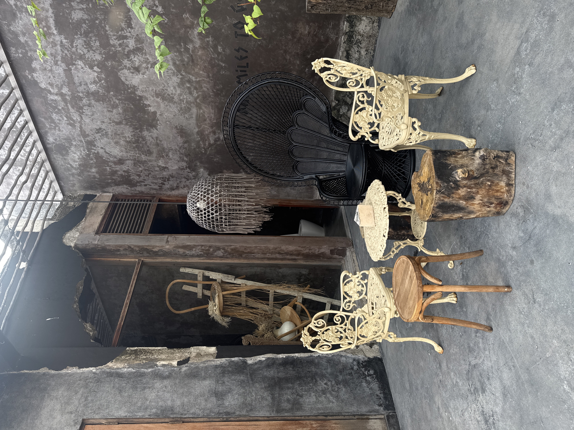 The image depicts an outdoor seating area in what appears to be a cafe or restaurant in Mengwi, Indonesia. The scene is furnished with a variety of seating options and decorative elements, creating a rustic and eclectic ambiance.
In the foreground, a circular, light-colored metal table with ornate detailing is positioned centrally. To its left, a small, round wooden stool with three legs sits on the concrete floor. To the right of the table, a rough-hewn tree trunk serves as a natural, rustic stool.
Two cream-colored metal chairs with intricate scrollwork and floral motifs flank the central table, one on each side. These chairs have a classic, vintage design. Behind the table and between the chairs, a large, black rattan peacock chair stands prominently. This chair is characterized by its high, fan-shaped back and woven texture.
In the background, a weathered, dark gray textured wall dominates. A wooden door is visible to the far left. To the right of the door, an opening leads into a darker interior space, where a white toilet can be seen. A wooden ladder leans against the wall within this opening, adorned with a straw hat and dried grasses, adding to the bohemian aesthetic. A white vase containing dried flowers is also present in this area.
Above the seating arrangement, a large, macrame-style pendant light hangs from the ceiling, its fringe cascading downwards. The ceiling itself appears to be constructed of wooden beams and possibly some translucent roofing material, allowing diffused light to enter the space.
On the far right wall, partially obscured by a green leafy branch, text is faintly visible, reading "MILES TO GO". The concrete floor is aged and shows signs of wear. The overall impression is of a curated, artistic outdoor space that blends vintage furniture with natural elements and a touch of industrial-chic.
