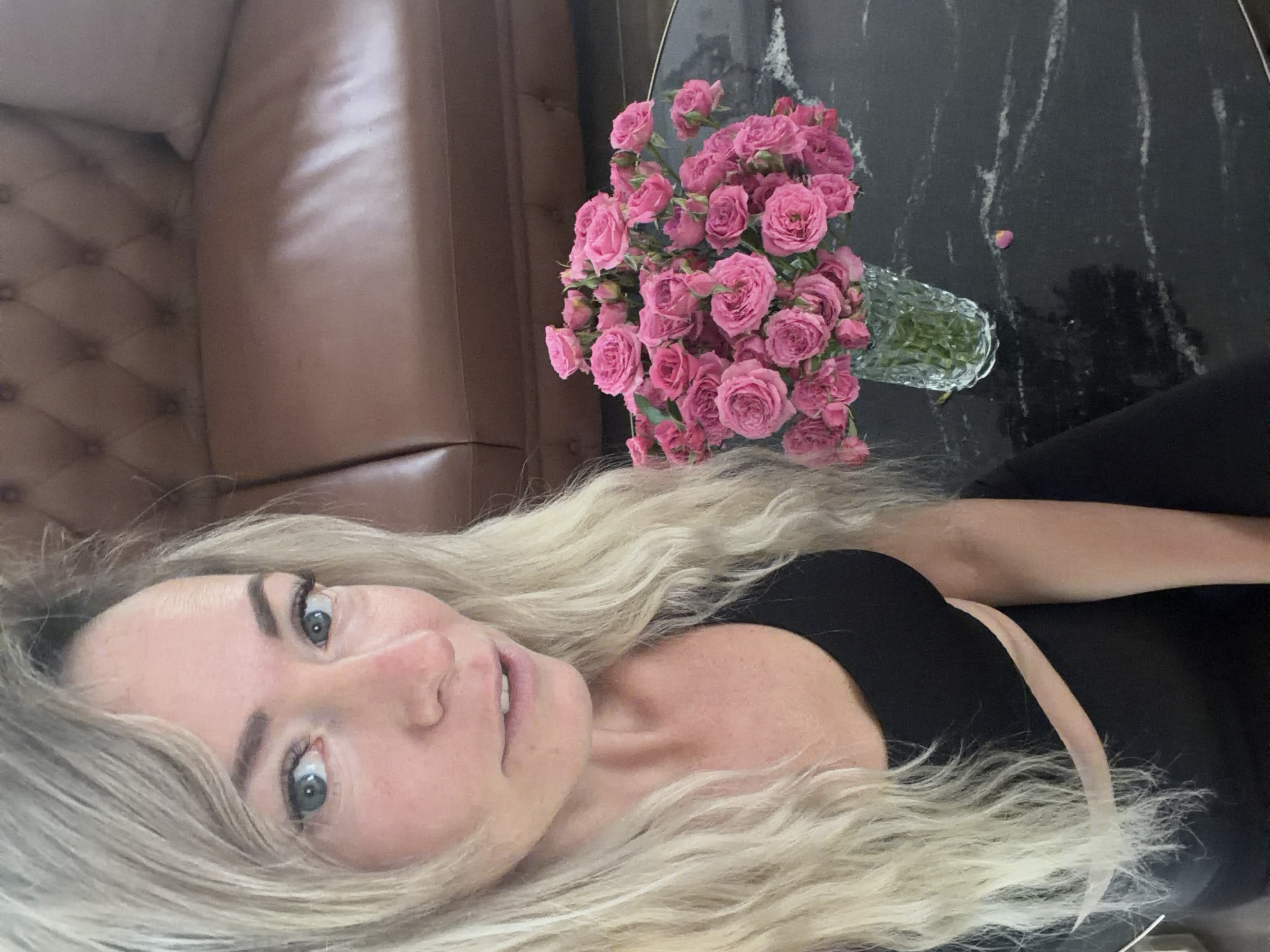 A selfie is taken by a blonde woman with blue eyes and fair skin, looking towards the camera. She is wearing a black top. To her right, on a dark marble table, is a clear glass vase filled with a bouquet of pink roses. In the background, a brown leather tufted couch is visible. The scene appears to be indoors, likely in a domestic setting.