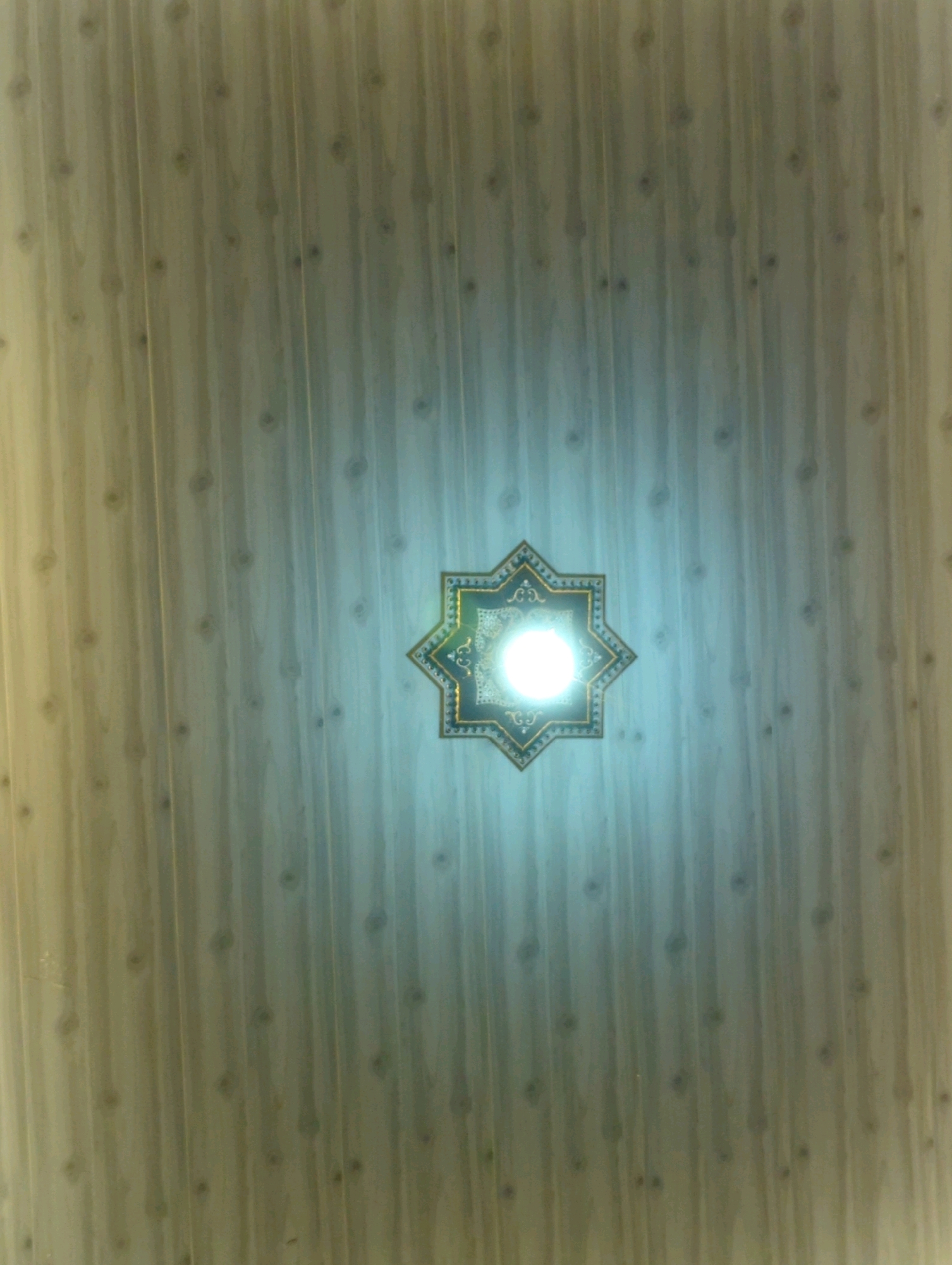 A low-angle, medium shot captures a ceiling in Kedungdowo, Indonesia, decorated with a repeating wood-grain pattern. In the center of the frame, a prominent eight-pointed star-shaped light fixture glows brightly. The fixture has a dark blue border with gold trim, and intricate white patterns fill the rest of its surface, surrounding the intensely lit center. The light emanating from the fixture casts a soft, diffused glow across the ceiling, highlighting the textured wood-grain and creating a subtle gradient of light from left to right. The overall impression is one of a well-lit interior space with a decorative focal point.