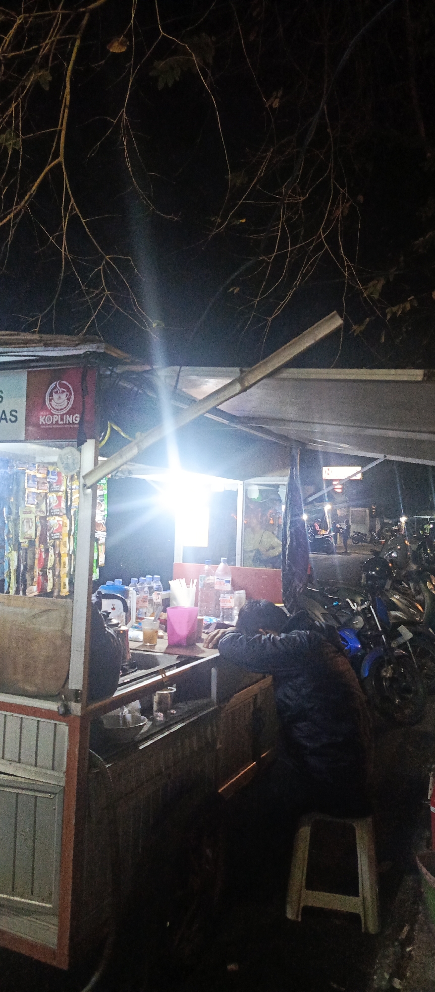 A dimly lit street-side food stall in Malang, Indonesia, is captured at night. The stall, constructed from wood and metal, is adorned with a "KOPLING" logo featuring a coffee cup, suggesting it sells beverages. Various items are displayed on shelves and counters, including packets of goods and bottled drinks. A bright light illuminates the scene from within the stall, creating a strong glare. In the foreground, a person wearing a dark jacket is seated at the counter, their head resting on their arms, appearing to be asleep or exhausted. To the right, several motorcycles are parked, indicating a common mode of transportation in the area. The background is dark, with tree branches visible against the night sky, and hints of other buildings or structures. The overall atmosphere is one of a quiet, late-night scene at a local food establishment.