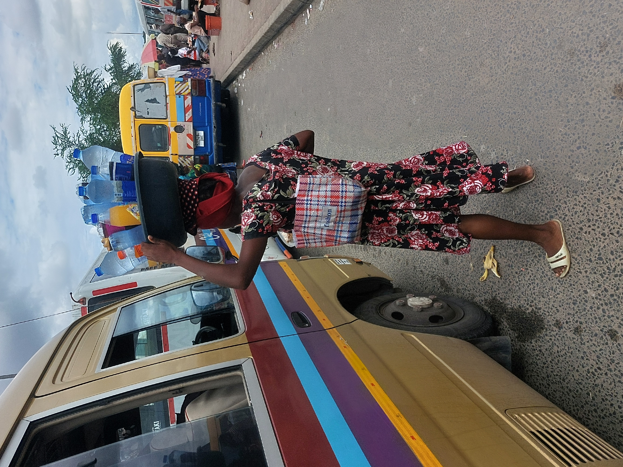 The image shows a woman in Dar es-Salaam, Tanzania, carrying a tray loaded with plastic bottles of water on her head, likely for sale. She is wearing a floral dress and sandals, and a red headscarf. She stands near a yellow minibus, which has colored stripes on its side. A banana peel lies on the ground near her feet. In the background, there is a crowd of people and other vehicles, suggesting a busy street scene.