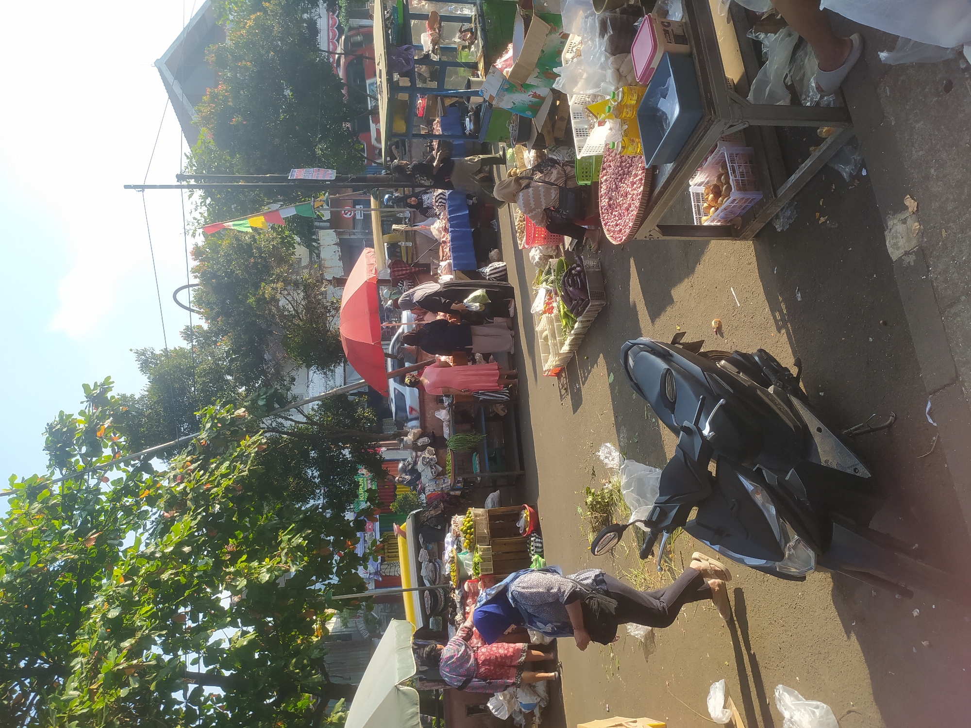 The image depicts a bustling outdoor market scene, likely in Kupang, Indonesia, given the presence of various Indonesian food items and the general ambiance of a local market. The time of day appears to be morning or early afternoon, suggested by the bright sunlight casting distinct shadows.
In the foreground, a dark-colored motorcycle is parked on the asphalt road, which is littered with some debris. To the left of the motorcycle, a woman wearing a blue hijab and a printed long-sleeved shirt is walking away from the camera, carrying a dark bag over her shoulder. She is wearing dark pants and light-colored sandals.
The market itself is organized with numerous stalls set up along the road. Several vendors are visible, interacting with customers or arranging their goods. One stall displays a variety of produce, including what appear to be oranges in crates, as well as leafy green vegetables. Another stall prominently features a large basket filled with what looks like shallots or red onions, alongside bottles of cooking oil and a pink container. Beneath this stall, a crate of eggs is visible. Further back, there are stalls with fabrics or textiles, and more food items like meat or poultry.
People are dressed in casual attire, with many women wearing headscarves (hijabs), typical of the region. A red umbrella is open at one of the stalls, providing shade. In the background, trees with green foliage line the market area, and a building with a tiled roof is visible. A few vehicles, including cars and what might be another motorcycle, are also present, indicating a busy thoroughfare. The overall impression is one of a lively, active marketplace where daily commerce is taking place.