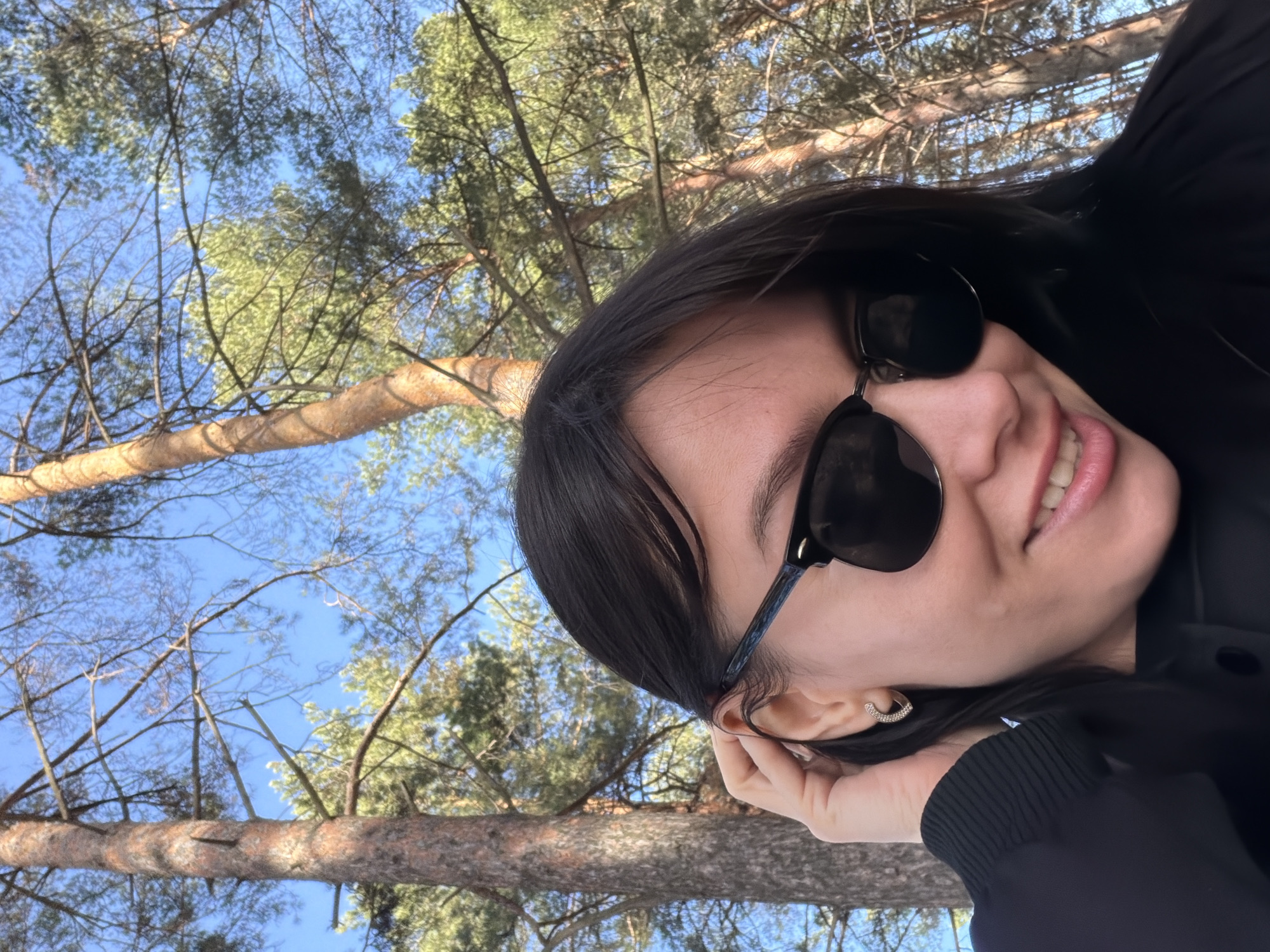 An outdoor, rotated selfie-style image captures a person smiling against a backdrop of pine trees and a clear blue sky. The photo is tilted 90 degrees clockwise, giving the impression of the subject lying down or leaning heavily to the side while looking up.
The main subject is a young person, likely female, with dark hair, wearing large, dark sunglasses and a black long-sleeved top or jacket. They are smiling broadly, revealing teeth, and have a hand resting near their right ear, where a small, golden hoop earring is visible. The skin tone appears warm.
In the background, numerous pine trees with slender trunks and branches covered in green needles fill the frame. A prominent tree trunk crosses horizontally through the mid-ground. The vibrant blue sky is visible between the tree branches, indicating bright, sunny weather. The light suggests it is daytime.
The setting appears to be a natural forested area, consistent with the provided location context of Chaikovsky, Russia, implying a pleasant and relaxed outdoor moment. There is no visible text in the image.