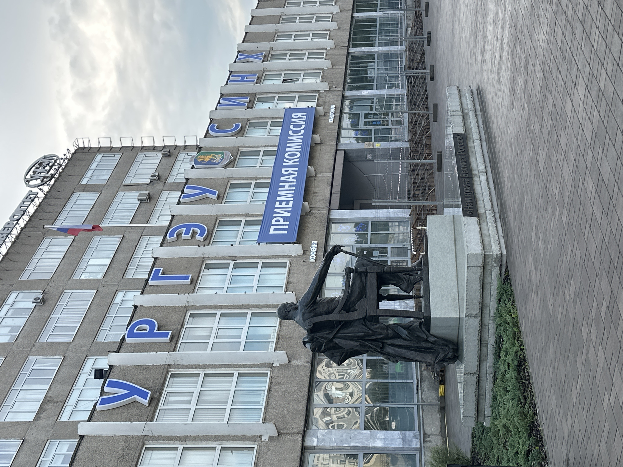 The image depicts a building with several signs and a sculpture in front of it. The building is tall and appears to be an office building or administrative center, with multiple stories and many windows. The facade is concrete and a bit weathered. On the upper floors, large blue letters are mounted, forming words that are partially visible. One prominent blue banner hangs vertically on the building, reading "ПРИЕМНАЯ КОМИССИЯ" (Reception Commission). To the left of this banner, a circular logo is visible, and further left, a Russian flag is attached to the building.
In front of the building, there is a dark bronze sculpture of two figures. One figure is seated at what appears to be a desk or table, with their arms outstretched as if writing or working. The other figure is positioned behind or next to the seated figure, also in a dynamic pose. The sculpture is set on a stone base. To the right of the sculpture, a paved walkway with a diamond pattern is visible, and to the left of the sculpture, there is a patch of green bushes.
Behind the sculpture and building, scaffolding suggests ongoing construction or renovation. The sky is overcast with clouds, indicating a possibly dull or cloudy day. The overall scene is urban, with a focus on the architectural features and the public art. The text visible includes "ПРИЕМНАЯ КОМИССИЯ" on the banner and other Cyrillic letters on the building. A small sign on the building near the scaffolding reads "КАЛИБРОВКА". At the base of the sculpture, an inscription is visible on the stone, partially obscured and difficult to read fully, but it appears to be in Cyrillic, possibly a dedication or motto. The time of day appears to be daytime, given the ambient light, but the cloudy sky limits distinct shadows. The scene evokes a sense of civic or institutional presence.