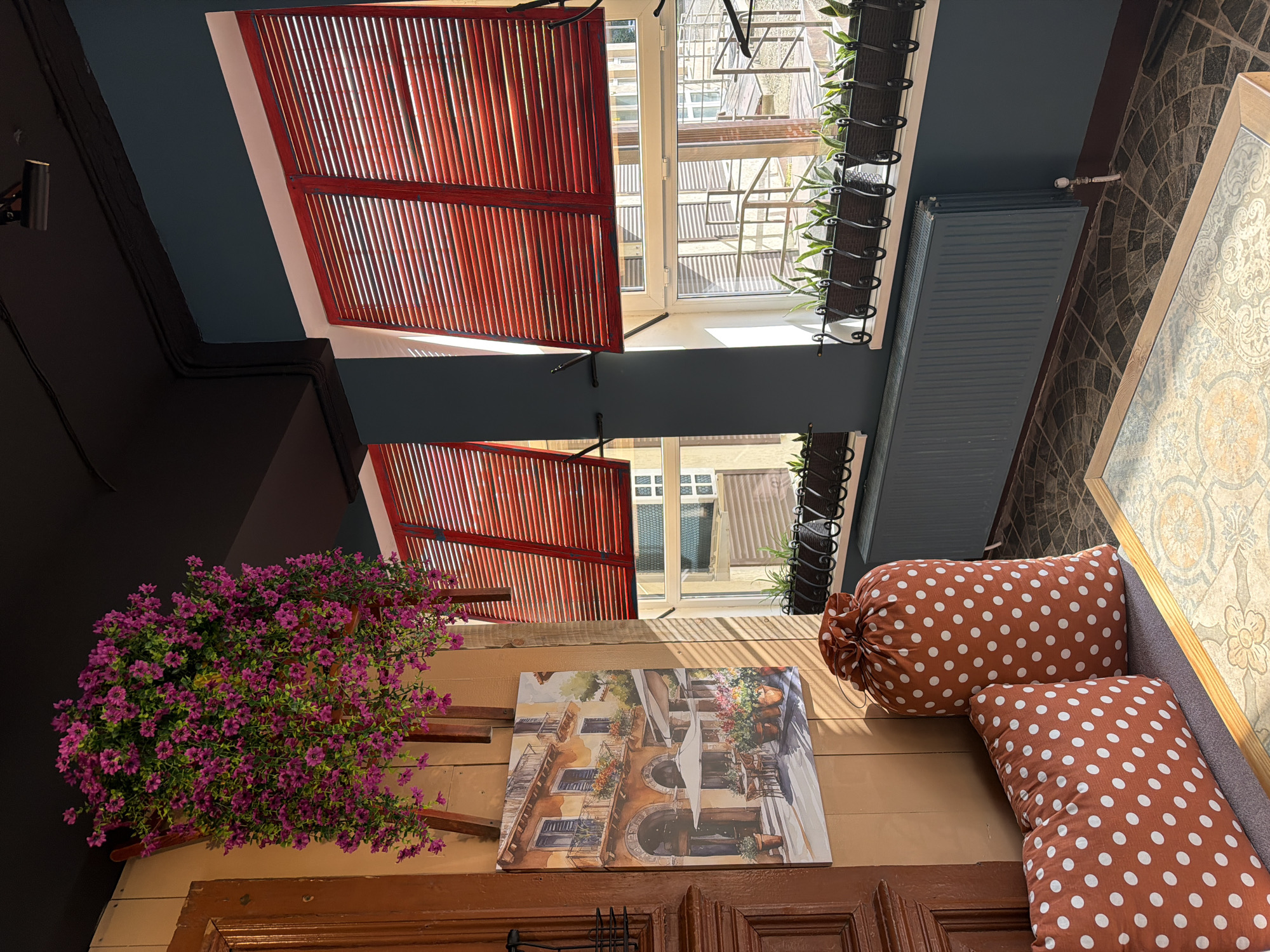 An upward-angled view captures an inviting, decorated space, likely a covered patio, balcony, or interior courtyard. The scene is dominated by architectural elements and decorative items.
Two sets of bright red louvered shutters are partially open, allowing natural light to stream in, creating strong vertical shadows. Beyond the shutters, a white window frame reveals a glimpse of an exterior building with various windows and structural elements. Below the upper set of shutters, a narrow black wrought-iron planter box filled with small green plants runs along a ledge.
The walls are painted in sections of dark grey-blue and a dark, possibly black, tone. A grey radiator is mounted on one of the dark grey-blue wall sections, with a white pipe connected above it. On the left side of the frame, a large, vibrant bush of artificial purple flowers spills downwards from a planter.
A horizontal wooden surface, possibly a wall or ceiling section, holds a framed painting depicting a European-style street scene with buildings, balconies, awnings, and potted flowers. Below this, a dark brown wooden door or panel is visible. To the right, two cozy brown cushions with white polka dots – one rectangular and one bolster-shaped – are placed on a grey seating area. The floor or a nearby surface features a patterned tile design.
The bright lighting and strong shadows suggest it is daytime, likely sunny. There are no people visible, and no specific activity is taking place. The setting evokes a sense of warmth and a blend of indoor and outdoor aesthetics. No visible text is present in the image.