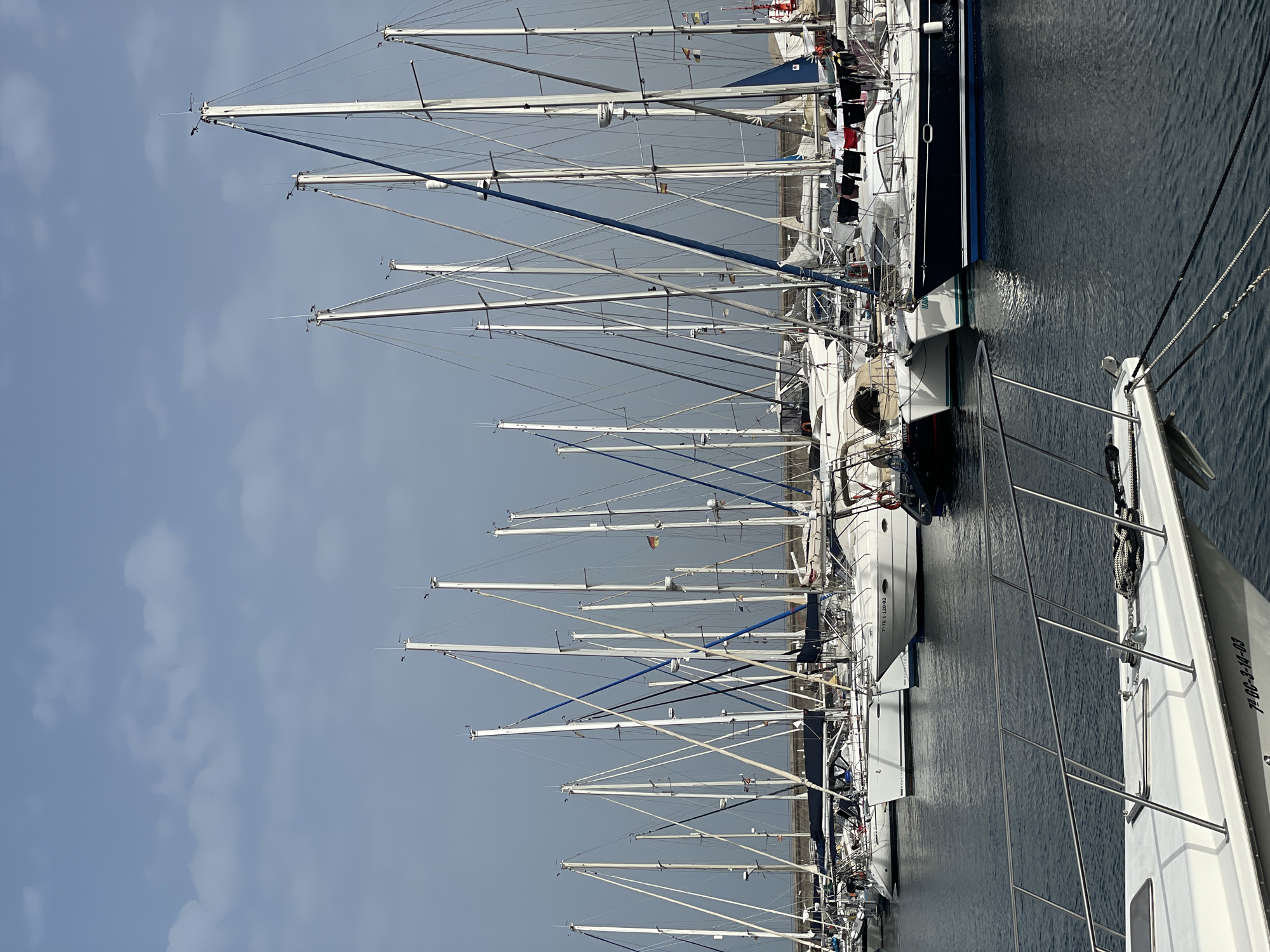 The image depicts a harbor filled with sailboats in Santa Cruz de Tenerife, Spain. Many masts are visible, ascending skyward. The water reflects the hulls of the boats. The sky is overcast with a grey-blue hue. Some masts are colored blue. Clothes are drying on one of the boat.