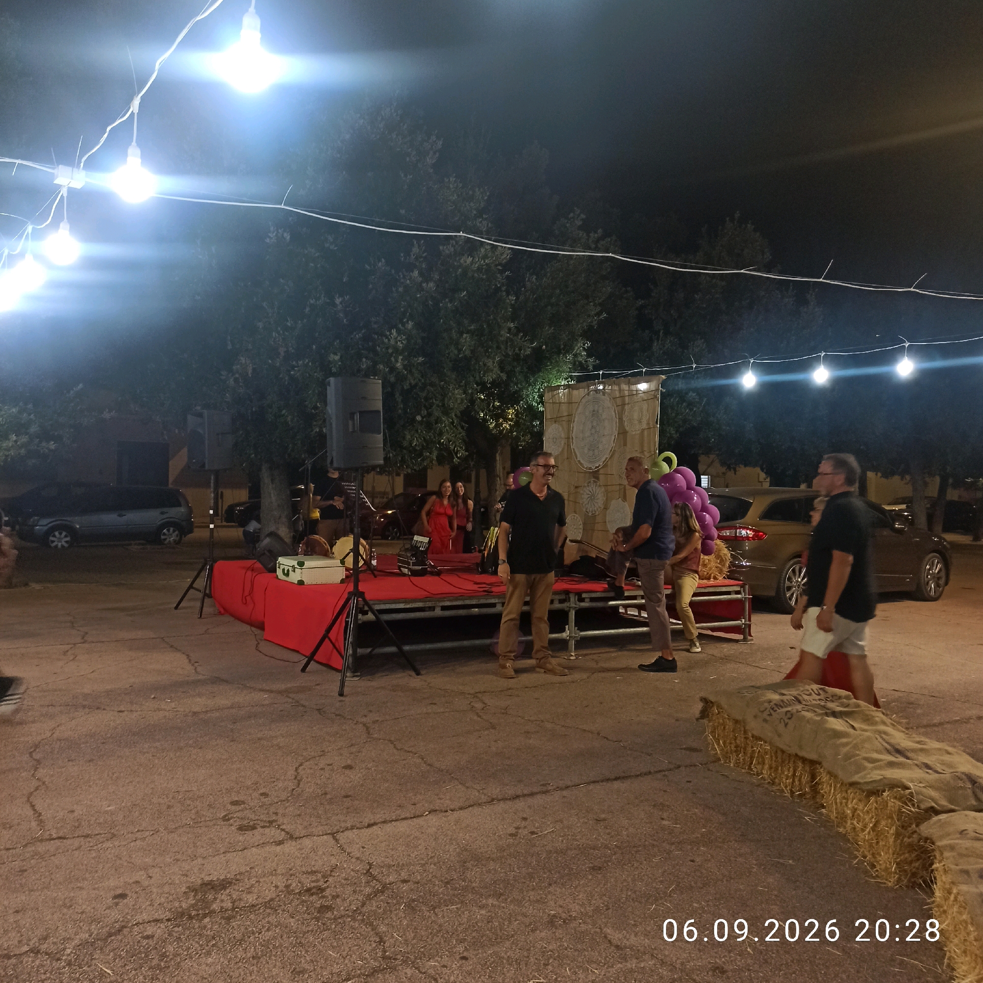 An outdoor nighttime scene features a raised stage with red carpeting, set up in what appears to be a town square or park in Bitonto, Italy. String lights with visible bulbs illuminate the area. Several people are present. On the stage, two men stand and converse. One man, wearing glasses and a dark polo shirt with tan pants, smiles. The other man, in a blue shirt and gray pants, stands behind him. A woman in a red dress is visible in the background on the stage. To the left of the stage, two large speakers on stands are positioned. In front of the stage, musical instruments, including what look like drums and an accordion, are set up on a table covered with a red cloth. A beige, hard-sided case rests on the table. In the background, trees with dense foliage are visible. Several cars are parked in the vicinity, including a dark-colored minivan and a gold sedan. In the foreground, bales of hay wrapped in burlap are arranged as seating or decoration. A date and time stamp, "06.09.2026 20:28", is visible in the lower right corner of the image.