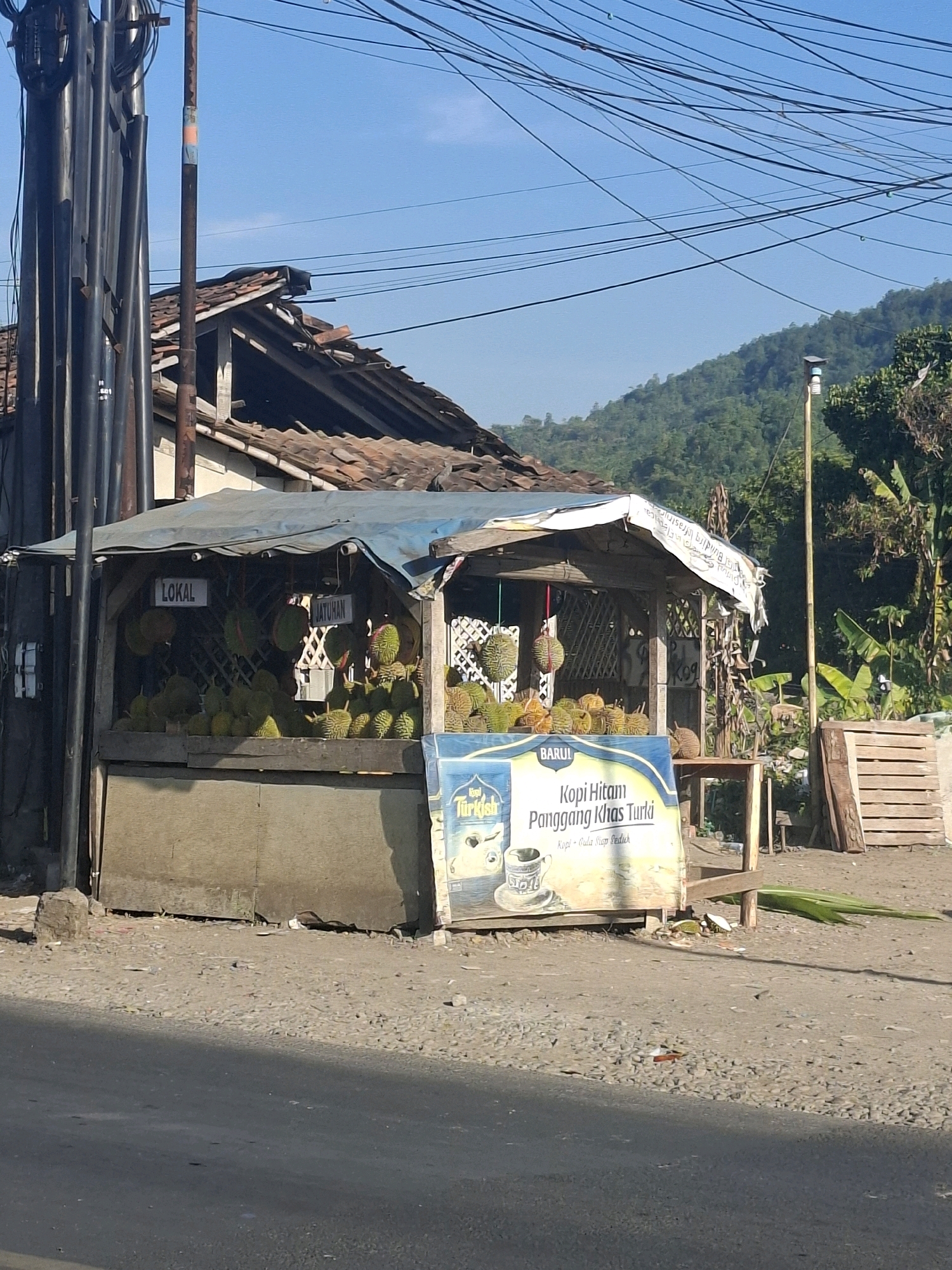 The image captures a roadside fruit stand in Lembursawah, Indonesia, under a clear blue sky. The stand, a simple structure with a blue tarp roof, is laden with a plentiful display of durians. Signs indicating "LOKAL" and "JATUHAN" are visible above the fruit. In front of the stand, a banner advertises "Kopi Hitam Panggeng Khas Turki," featuring an illustration of a coffee cup.
To the left of the stand, a tall utility pole with numerous wires is prominent. Behind the stand, an old, dilapidated building with a broken tile roof stands partially visible. To the right, a stack of wooden pallets and lush green vegetation, including banana trees, suggest a rural or semi-rural setting. The ground is covered with gravel and dirt, with a paved road in the foreground. The overall scene depicts a typical roadside vendor scene in Indonesia, likely focused on the sale of local produce and possibly coffee. There are no people visible in the frame.