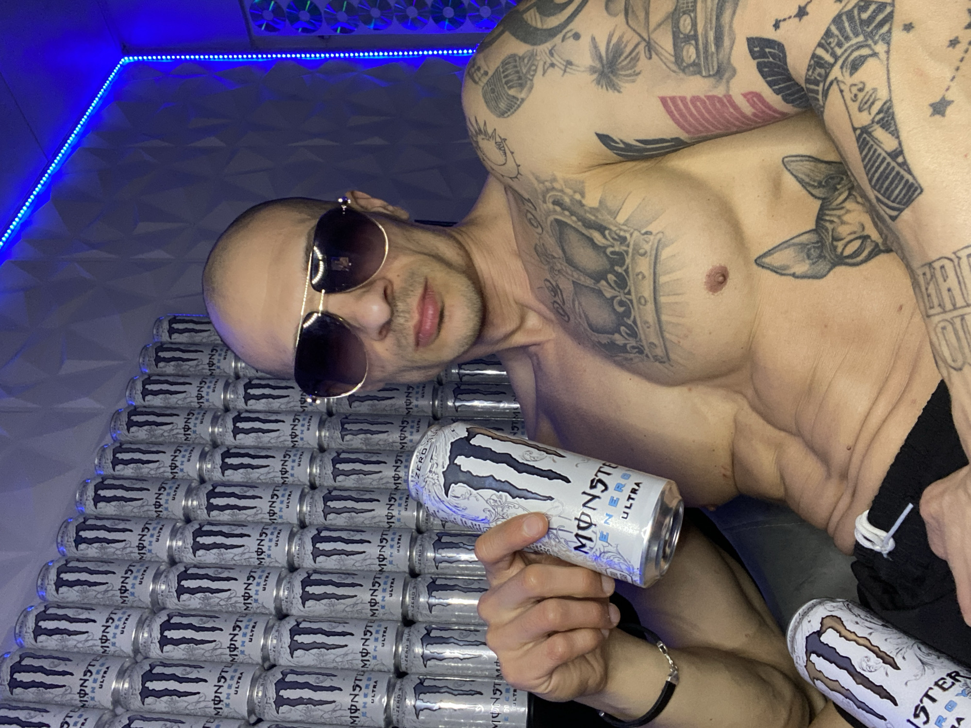 A bald, lightly tanned man wearing sunglasses poses shirtless in a room illuminated by blue LED strip lighting. He is surrounded by stacked cans of Monster Energy Ultra drink, both on a surface beside him and held in his hands. The man is heavily tattooed, with visible designs on his chest, shoulders, and arms. The background features a textured geometric pattern. The exact location in the city of Pleven, Bulgaria, is not discernible from the image.