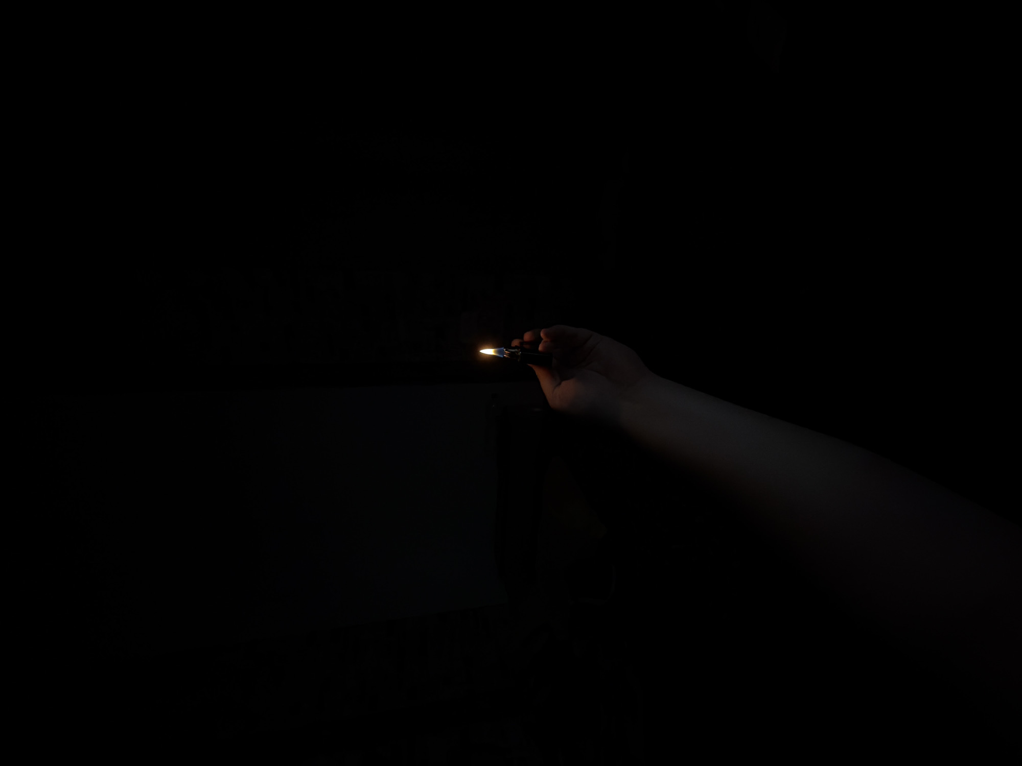 A hand holds a lit lighter in the dark. The lighter is black and has a flame that emits a warm glow. The hand is pale and appears to be a person's right hand. The background is completely black, creating a stark contrast with the illuminated lighter. The lighting suggests the image was taken in a dimly lit environment.